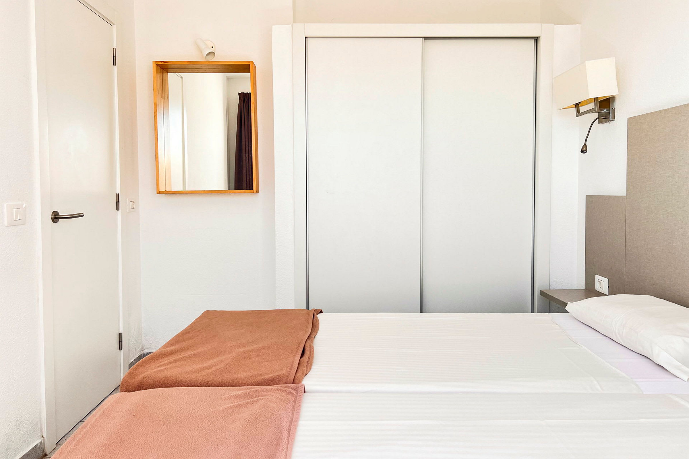 Ett hotellrum med vit inredning, en dubbel säng med vita lakan och bruna överkast, samt en spegel på väggen. (AI-genererad text)