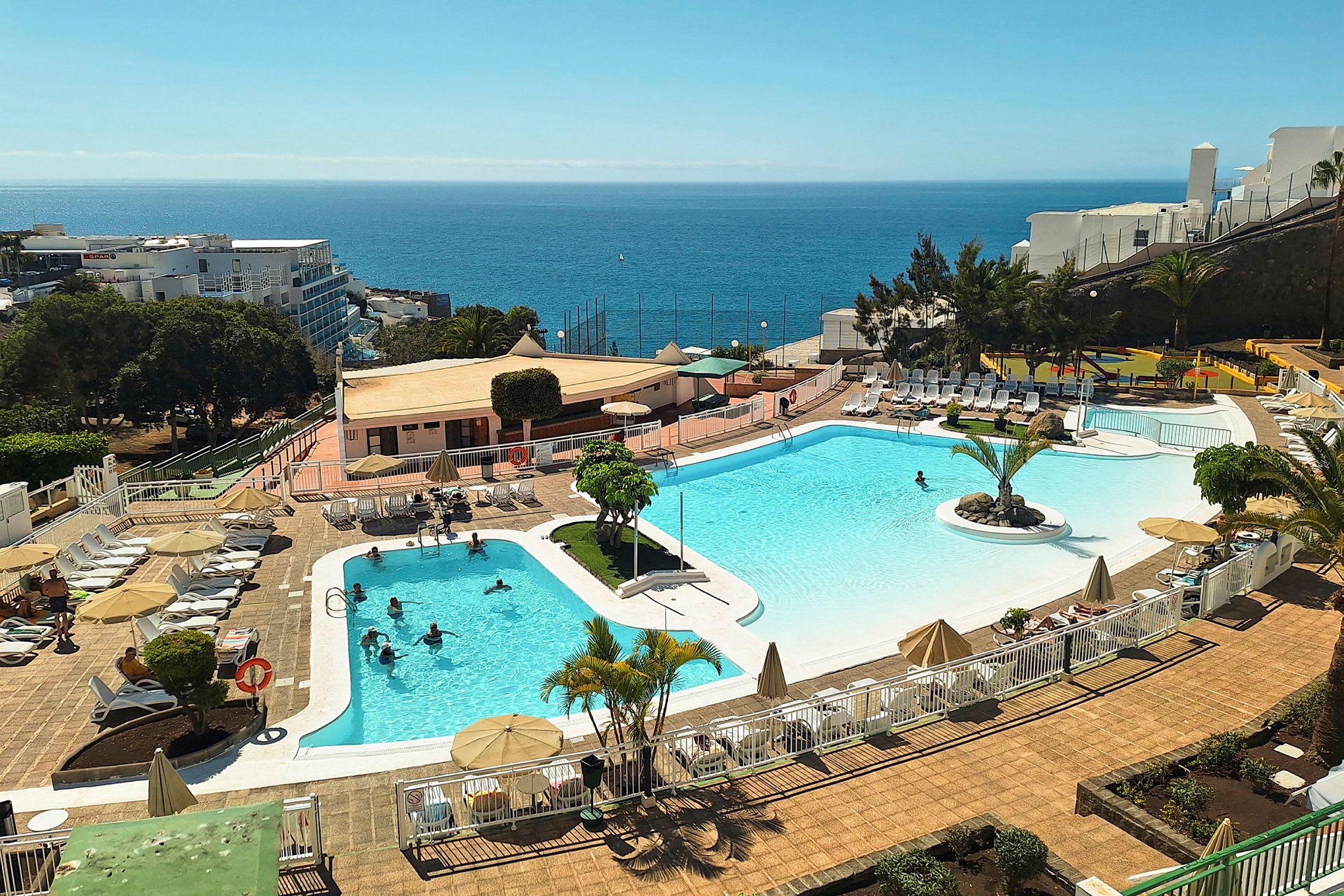 En utomhuspool vid ett semesterhotell med solstolar och parasoller. Havet syns i bakgrunden. (AI-genererad text)