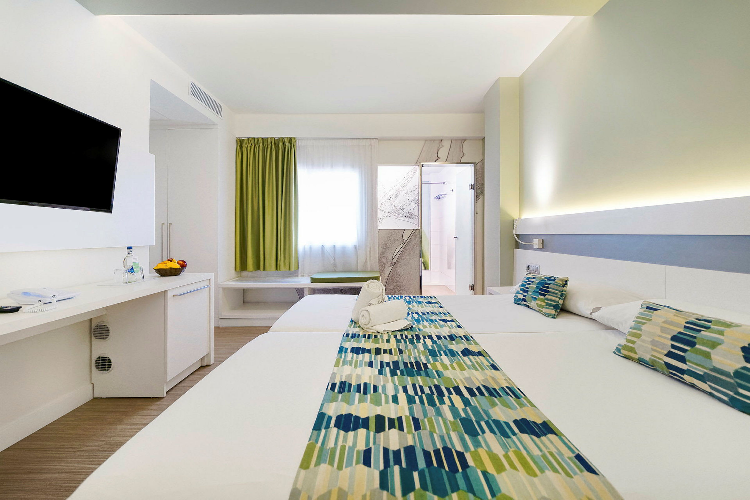 Ett hotellrum med två sängar, en TV på väggen, och en dörr till badrummet. Rummet har ljus inredning. (AI-genererad text)