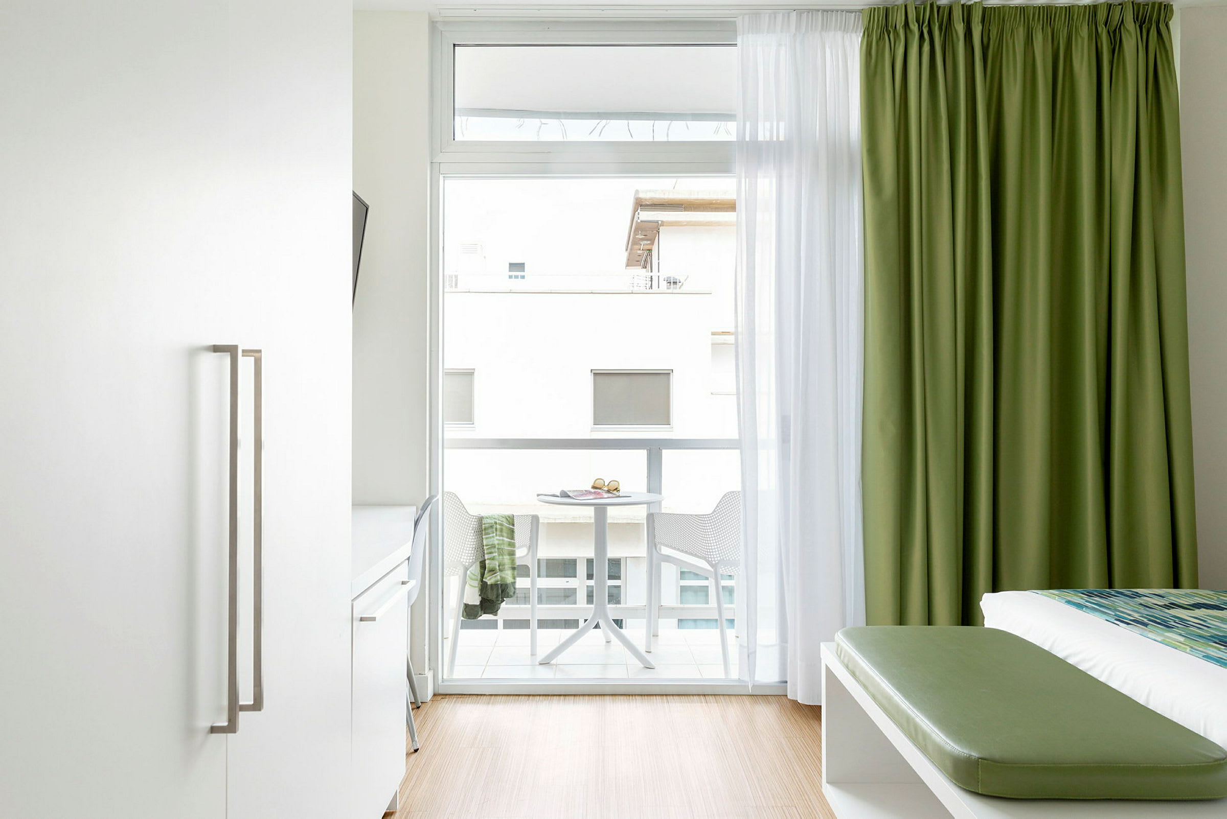 En ljus hotellrumsinteriör med en säng, gröna gardiner och en balkong med bord och stolar. (AI-genererad text)