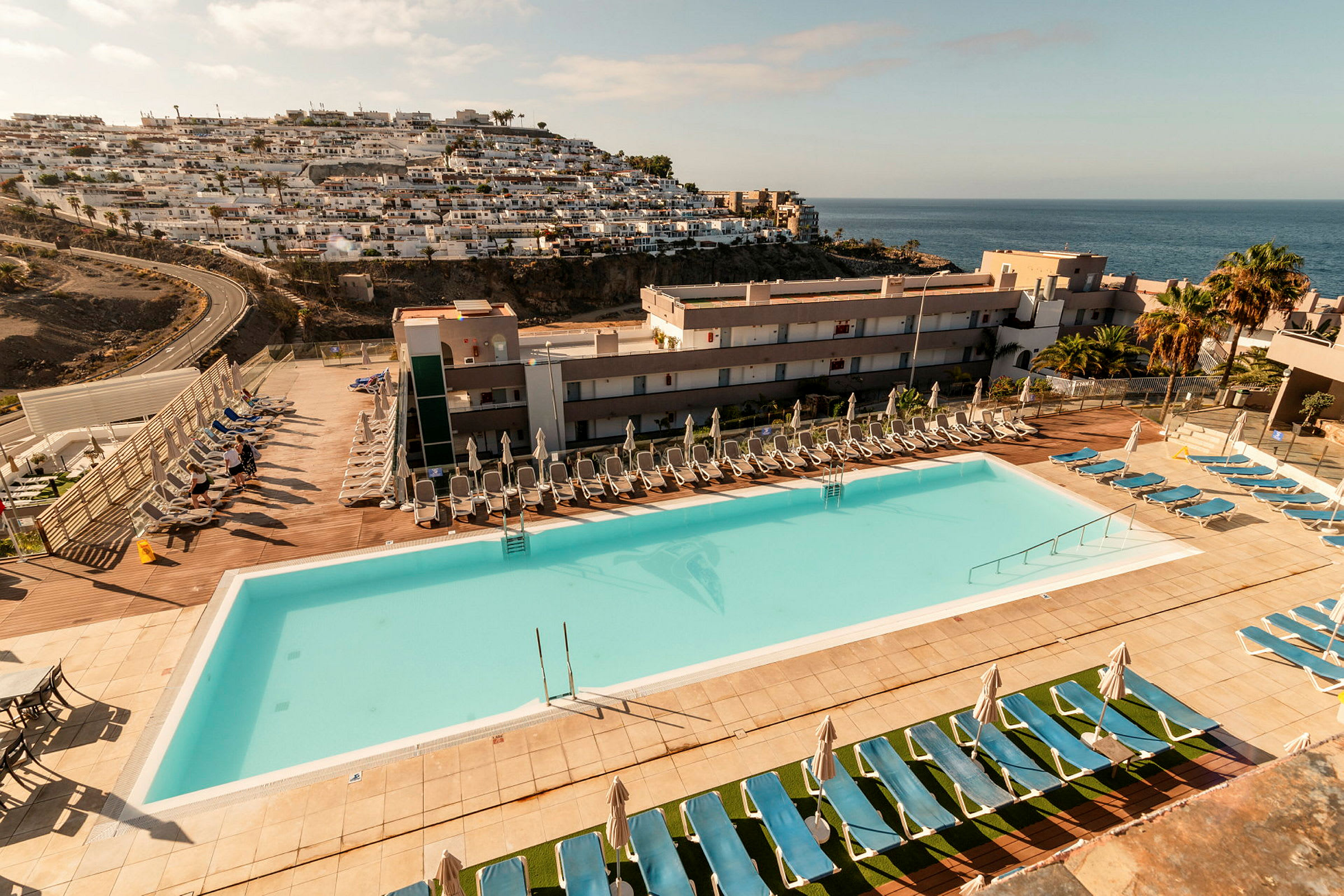 En utomhuspool med solstolar och parasoller vid ett hotellområde, med havsutsikt och bergssluttning med byggnader. (AI-genererad text)