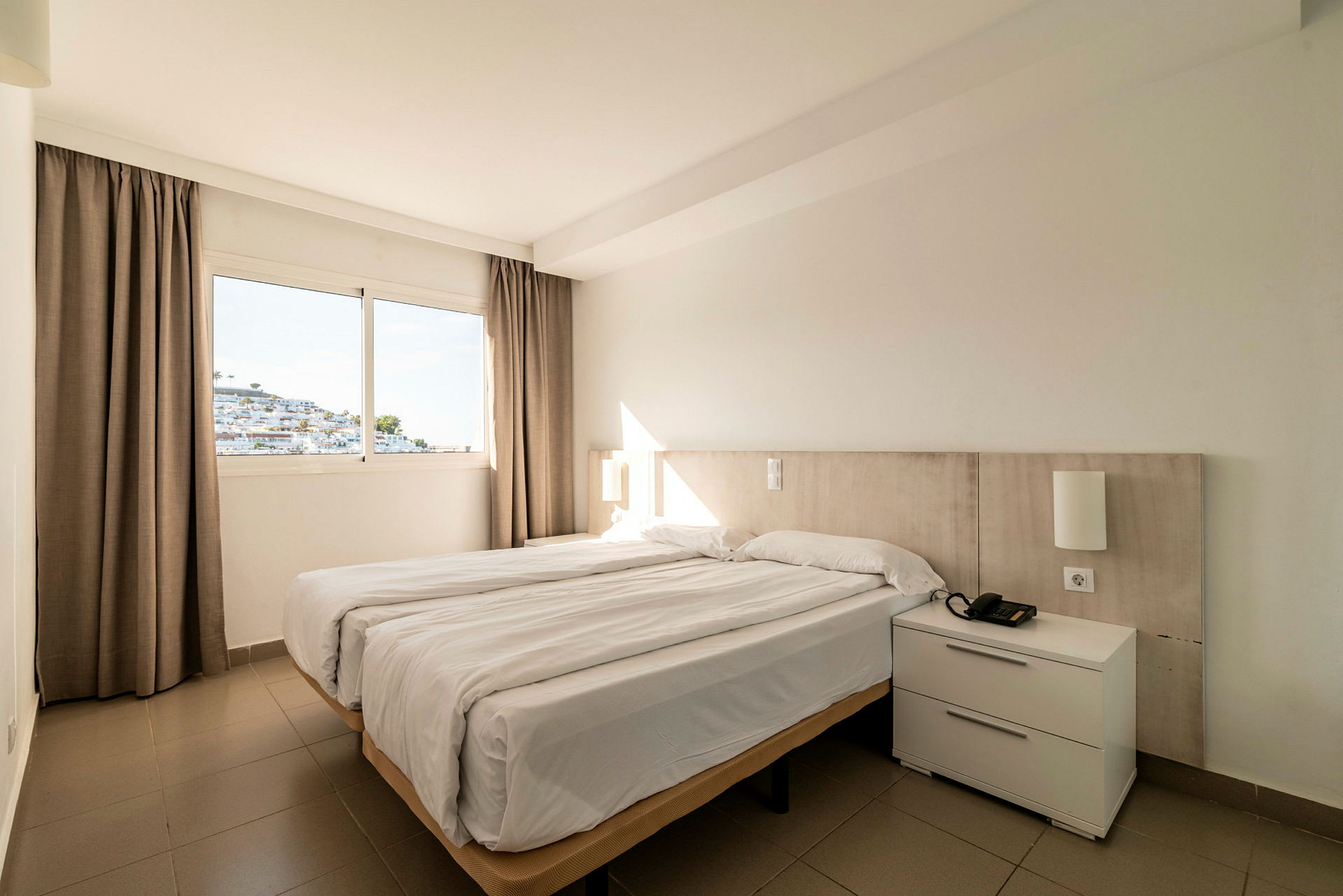 Ett hotellrum med en dubbelsäng och ett fönster som visar en vit byggnad. (AI-genererad text)