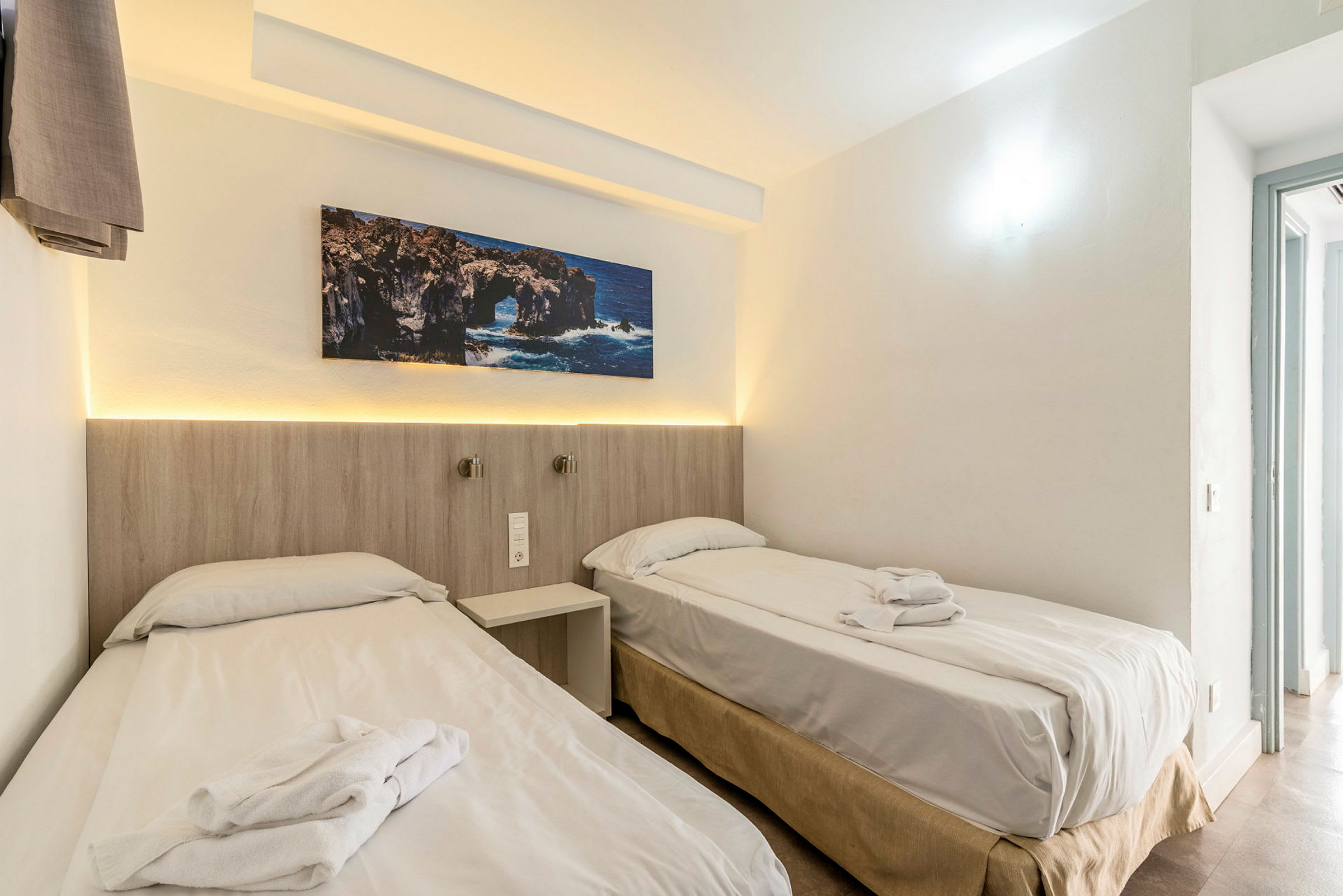 Två enkelsängar med vita lakan och handdukar på en ljus hotellrum vägg, med en klippmålning ovanför. (AI-genererad text)