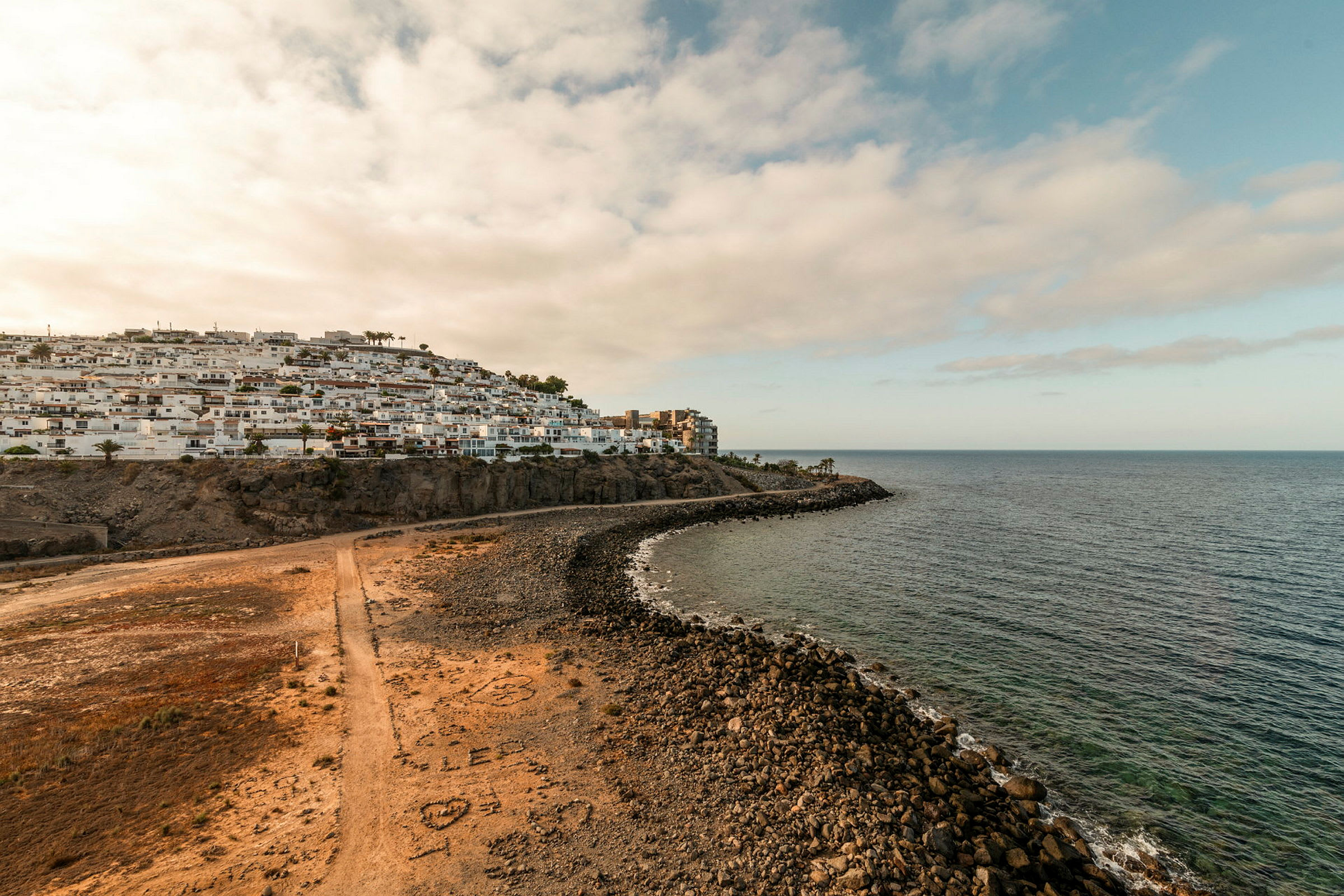 En kust med klippor, strand och hav med byggnader på en höjd vid sidan. (AI-genererad text)