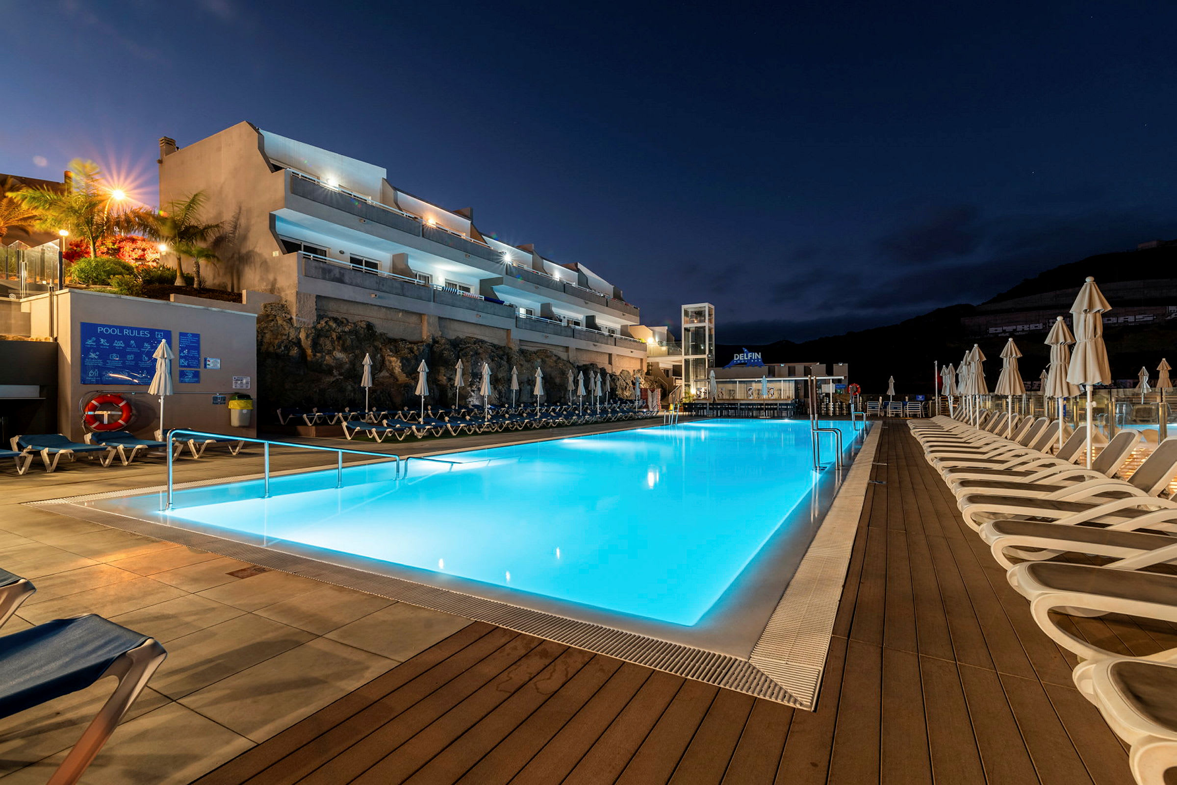 Exteriör vy av en upplyst swimmingpool vid natt tid, omgiven av solstolar och byggnader i bakgrunden. (AI-genererad text)
