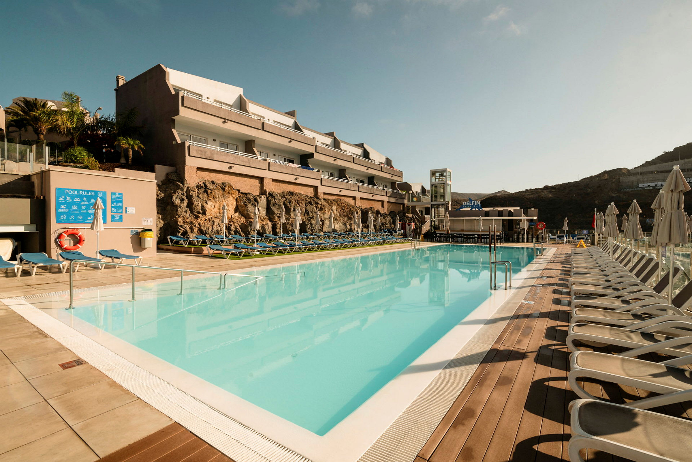 Utomhuspool med solstolar och hotellbyggnad i bakgrunden. (AI-genererad text)