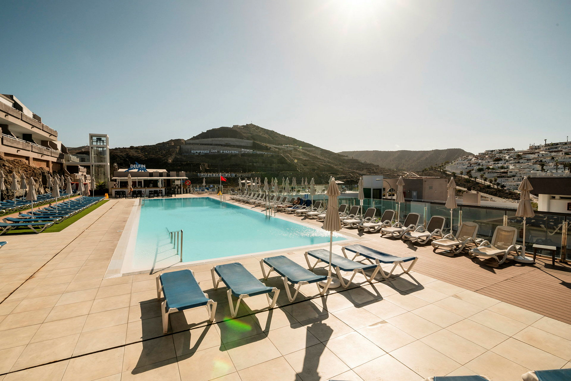 Exteriör vy av en hotellpool med solstolar och paraplyer på en solig bergig plats. (AI-genererad text)