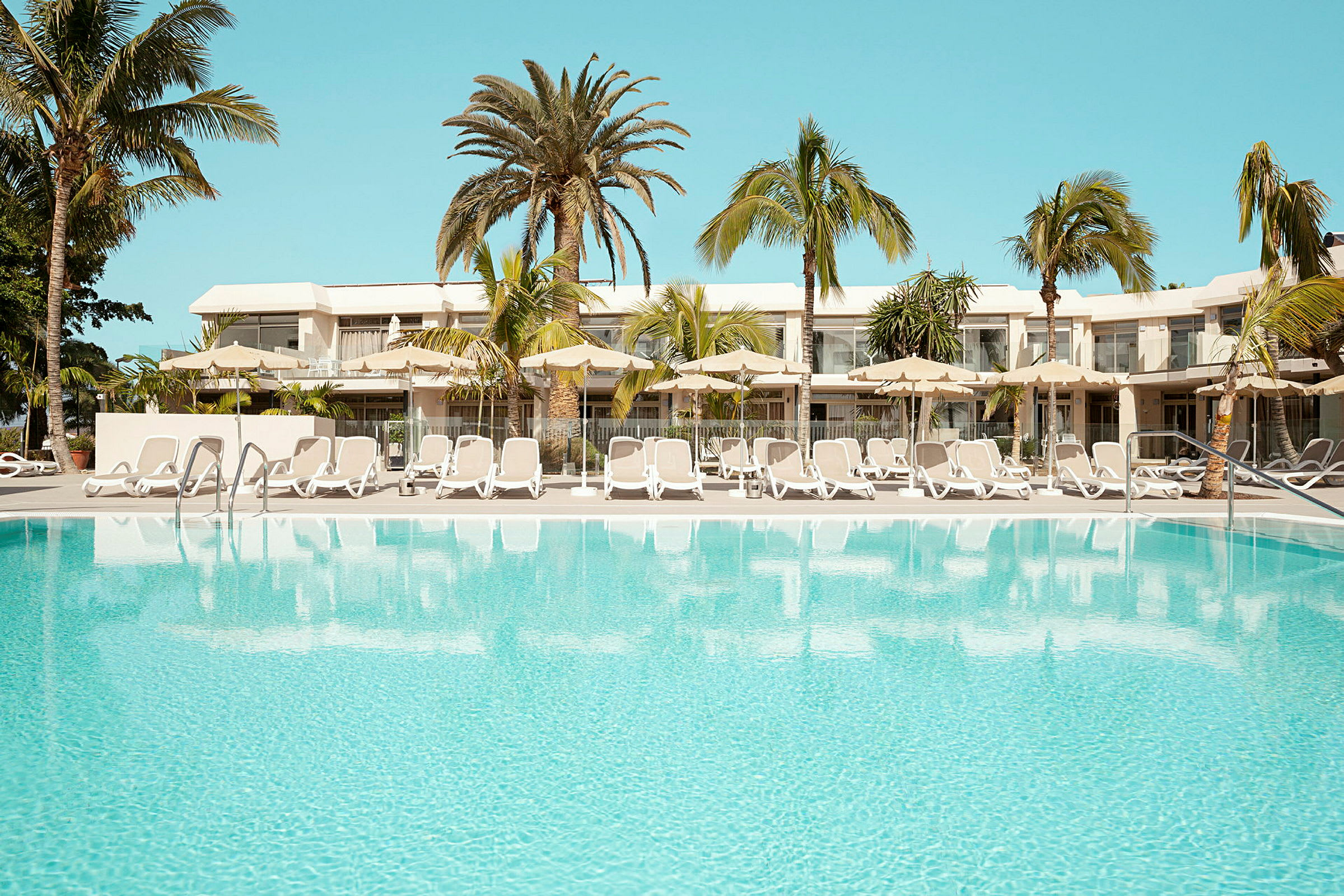 Utomhuspool omgiven av solstolar och parasoller, med ett hotell och palmer i bakgrunden. (AI-genererad text)