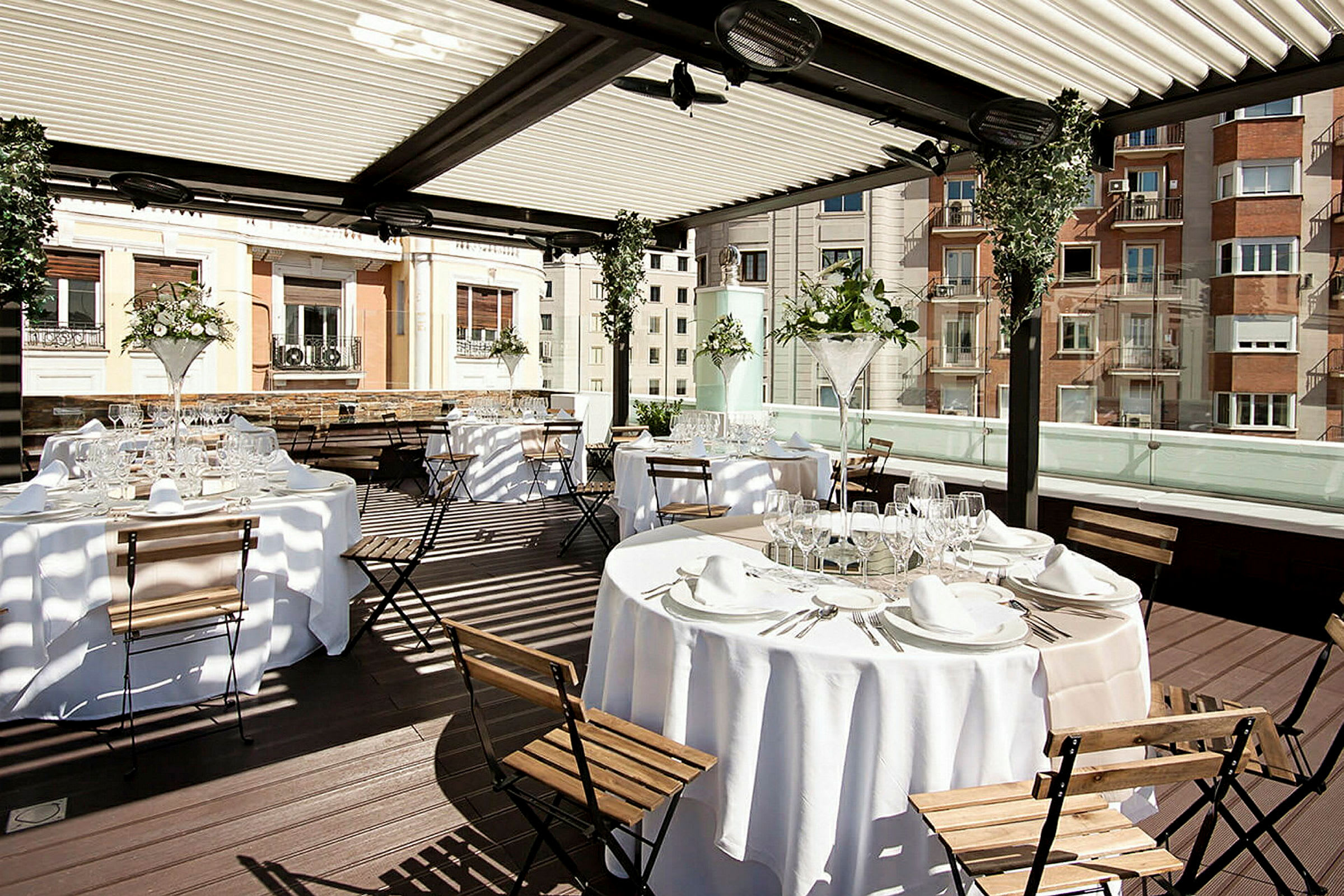 Bilden visar ett elegant utomhusområde med dukade bord och trästolar under en täckt pergola. (AI-genererad text)