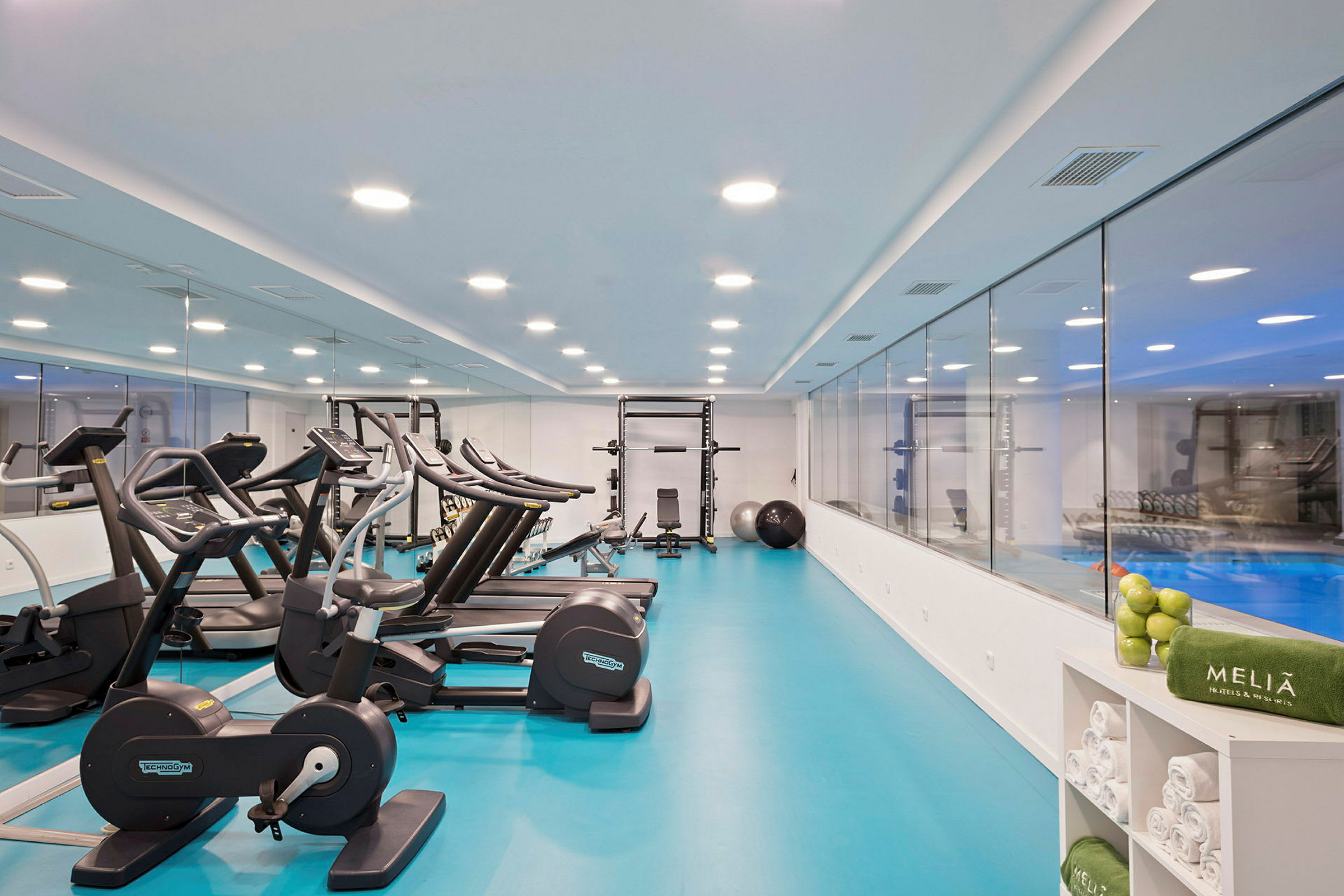 Ett modernt gym med träningsmaskiner, inklusive löpband och motionscyklar, samt en spegelvägg och en glasvägg som vetter mot en pool. (AI-genererad text)