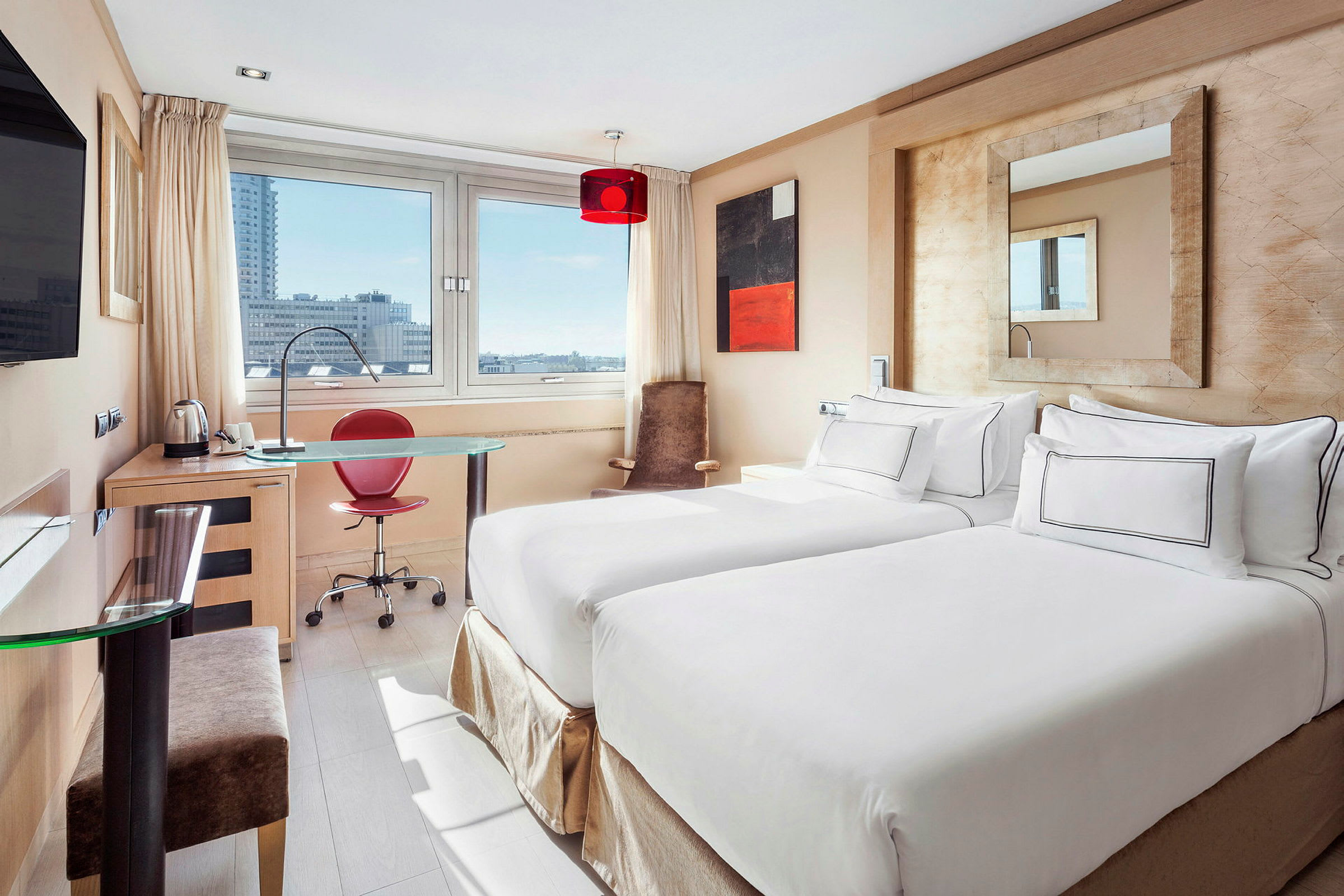 Ett hotellrum med två enkelsängar, ett skrivbord och en stor fönstermiljö som visar stadsutsikt. (AI-genererad text)