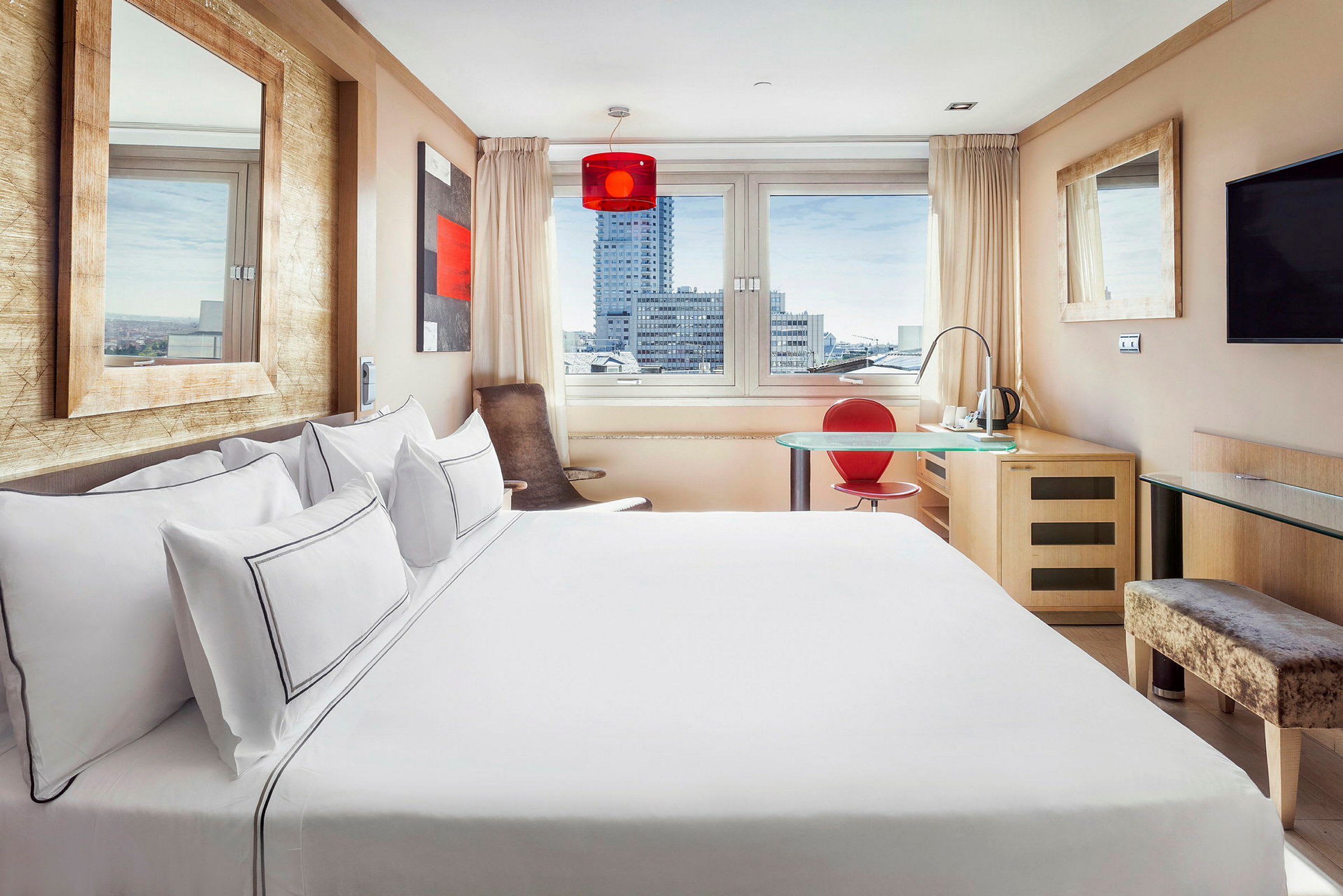 Ett ljus och modernt hotellrum med stor säng, fönster med utsikt över stadens byggnader, ett skrivbord med röd stol och en väggmonterad TV. (AI-genererad text)