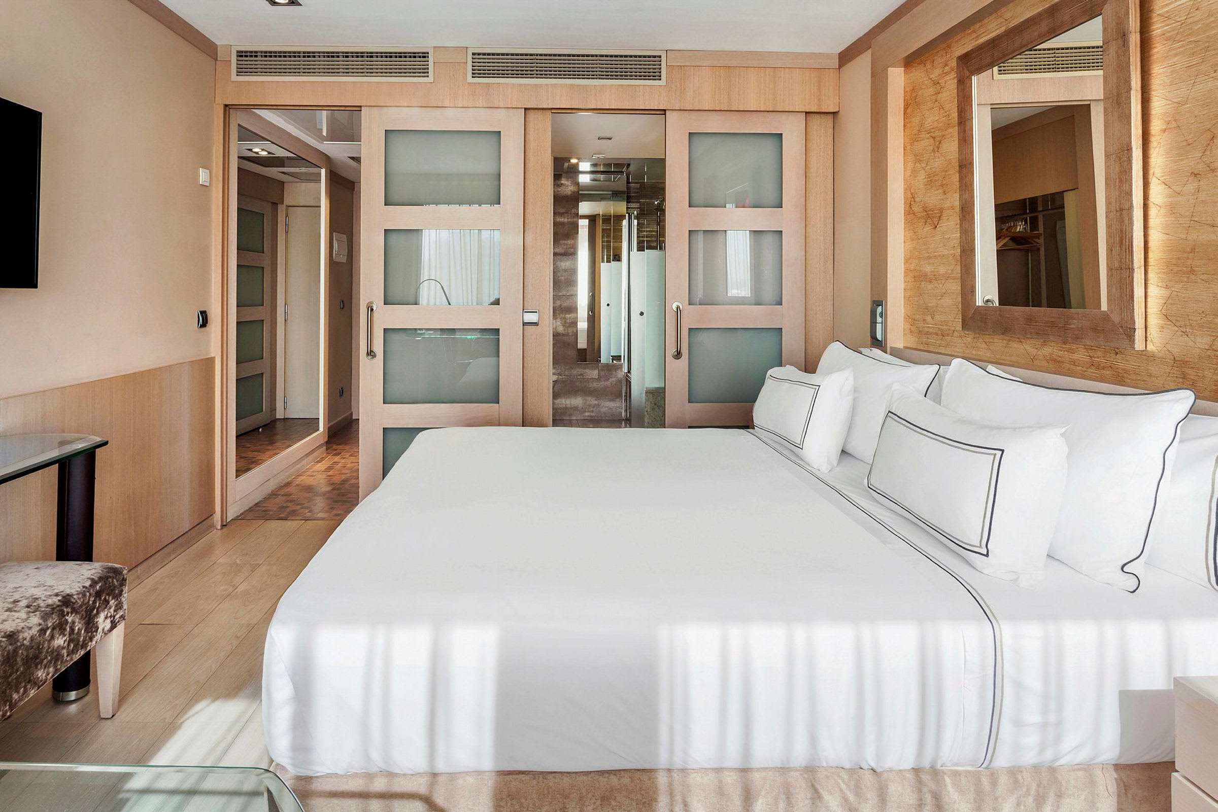 Ett modernt hotellrum med en stor säng klädd i vita sängkläder, en spegel på väggen, och en skjutdörr som leder till ett badrum. (AI-genererad text)
