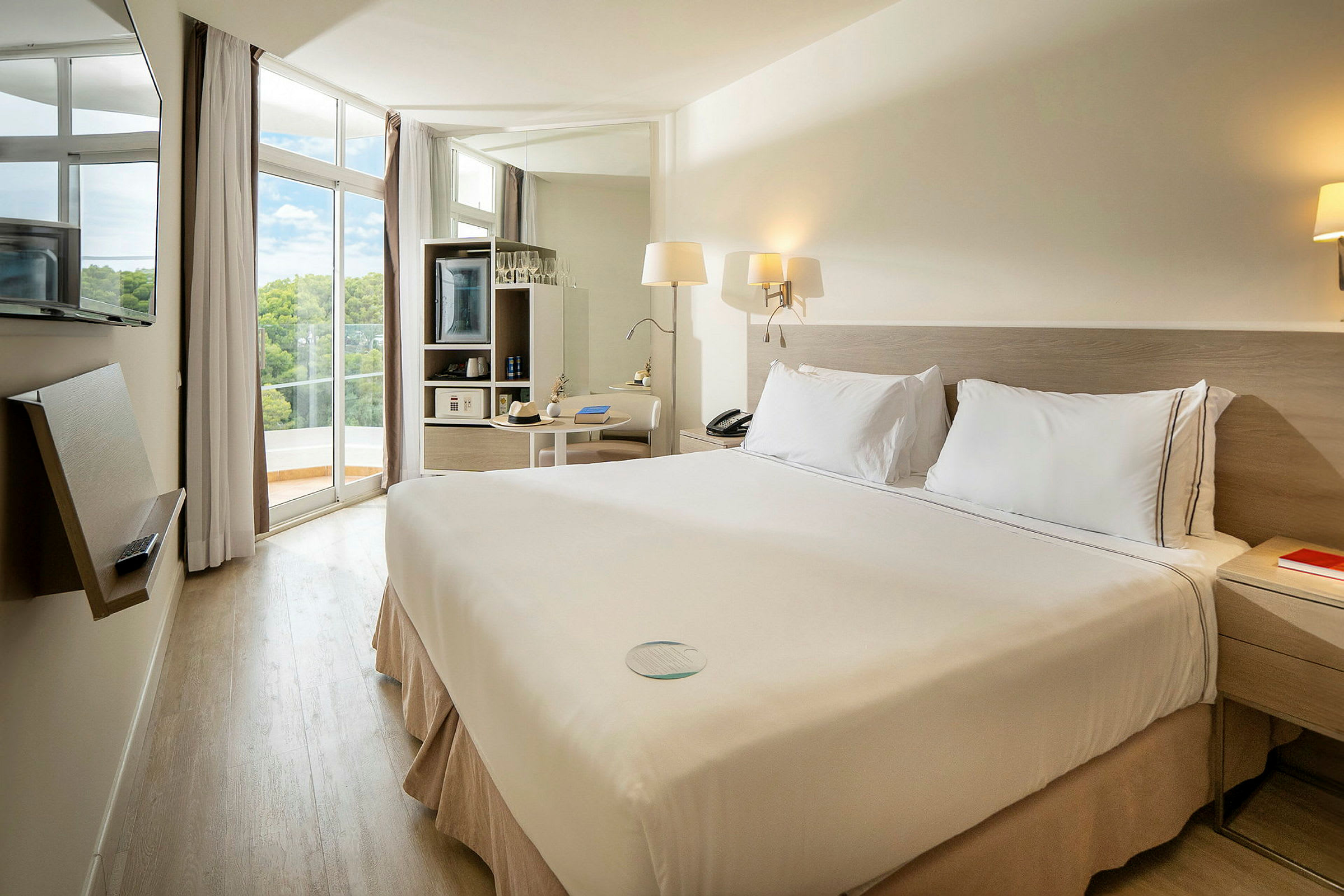 Hotelrum med dubbelsäng och ljus inredning, stora fönster och balkong med utsikt. (AI-genererad text)