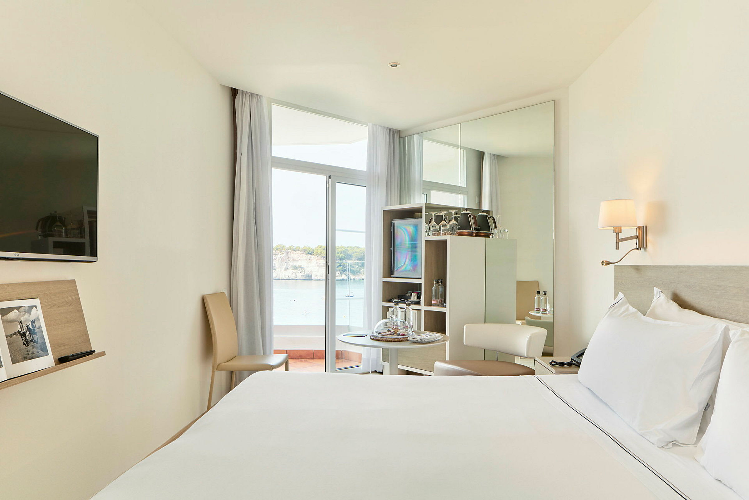 Ett hotellrum med en stor säng, ett litet bord med stolar, och en utsikt mot havet genom en glasdörr. (AI-genererad text)
