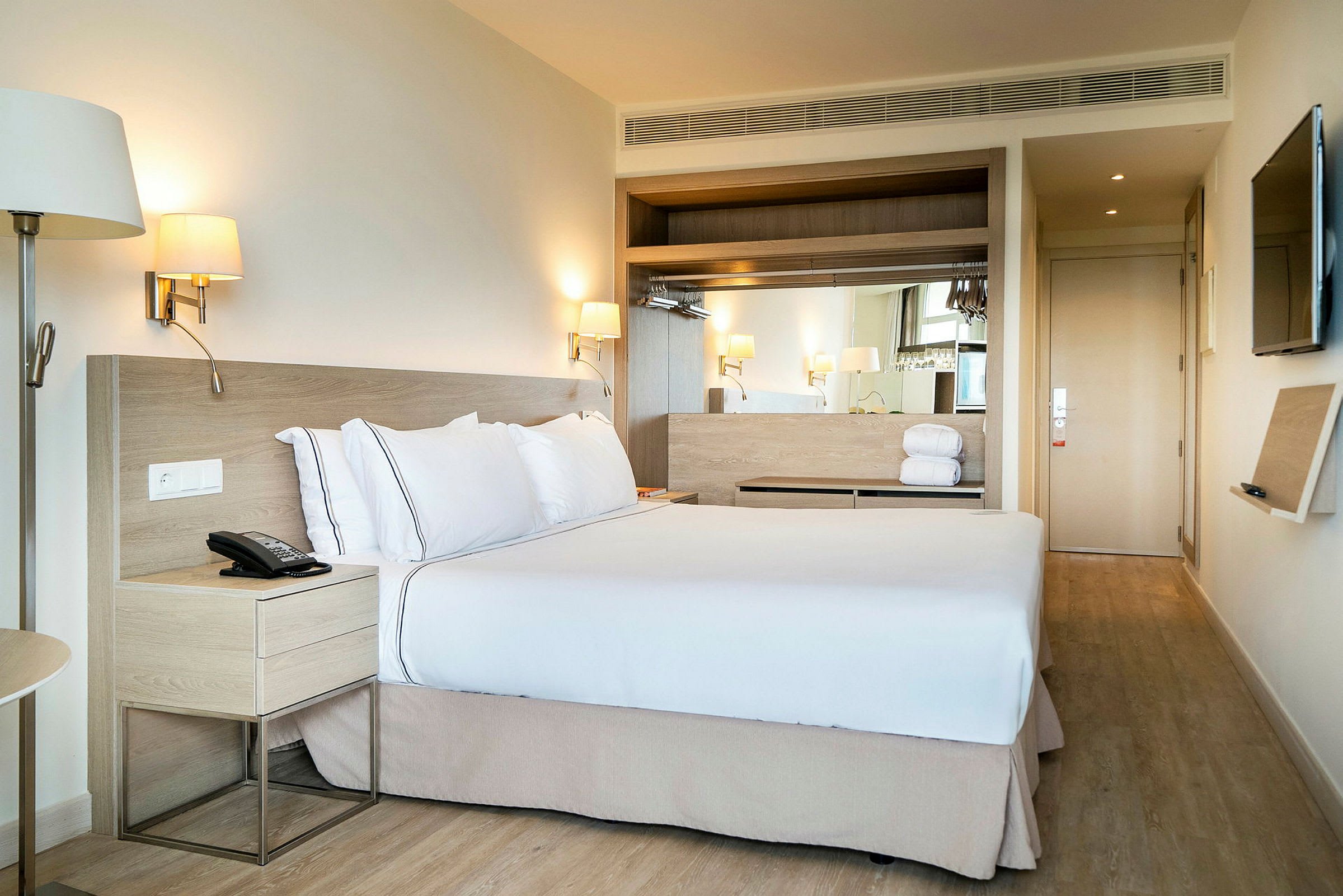 Ett hotellrum med en stor säng bäddad med vita lakan och kuddar. Lampor på väggen ovanför sängens sidor samt ett nattduksbord bredvid. (AI-genererad text)
