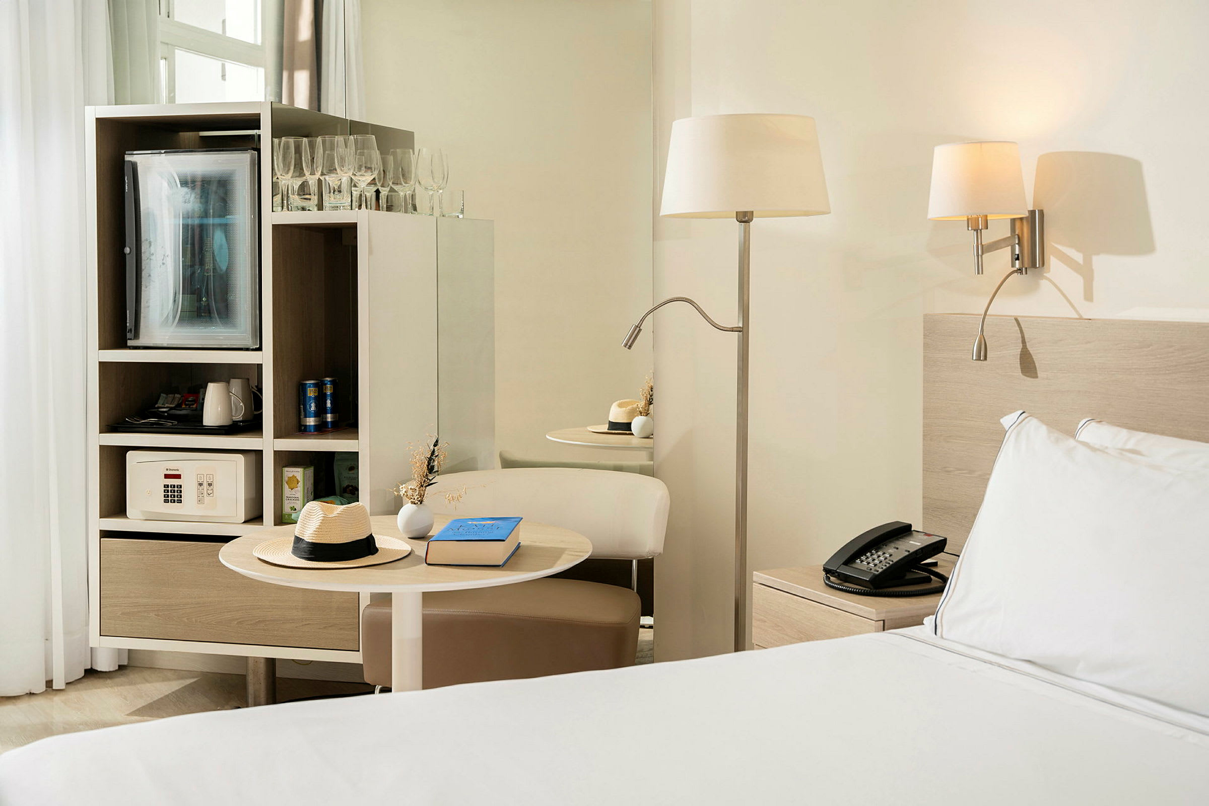 Ett hotellrum med en säng bredvid ett nattduksbord, en bordslampa och en inbyggd möbel som innehåller ett kylskåp och ett kassaskåp. (AI-genererad text)