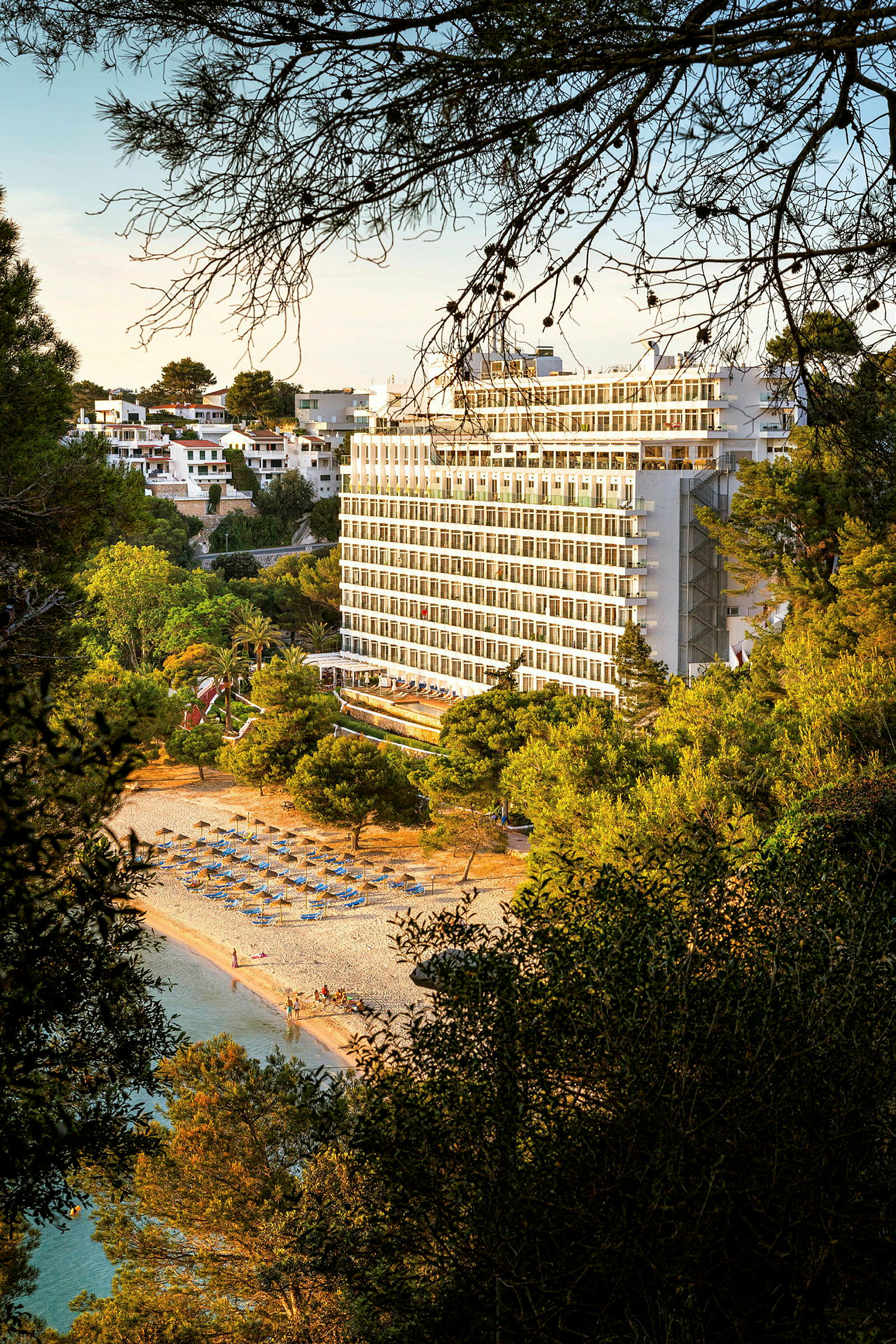 Bild av ett hotell byggt nära en strand med solstolar och parasoller, omgiven av grönskande träd. (AI-genererad text)