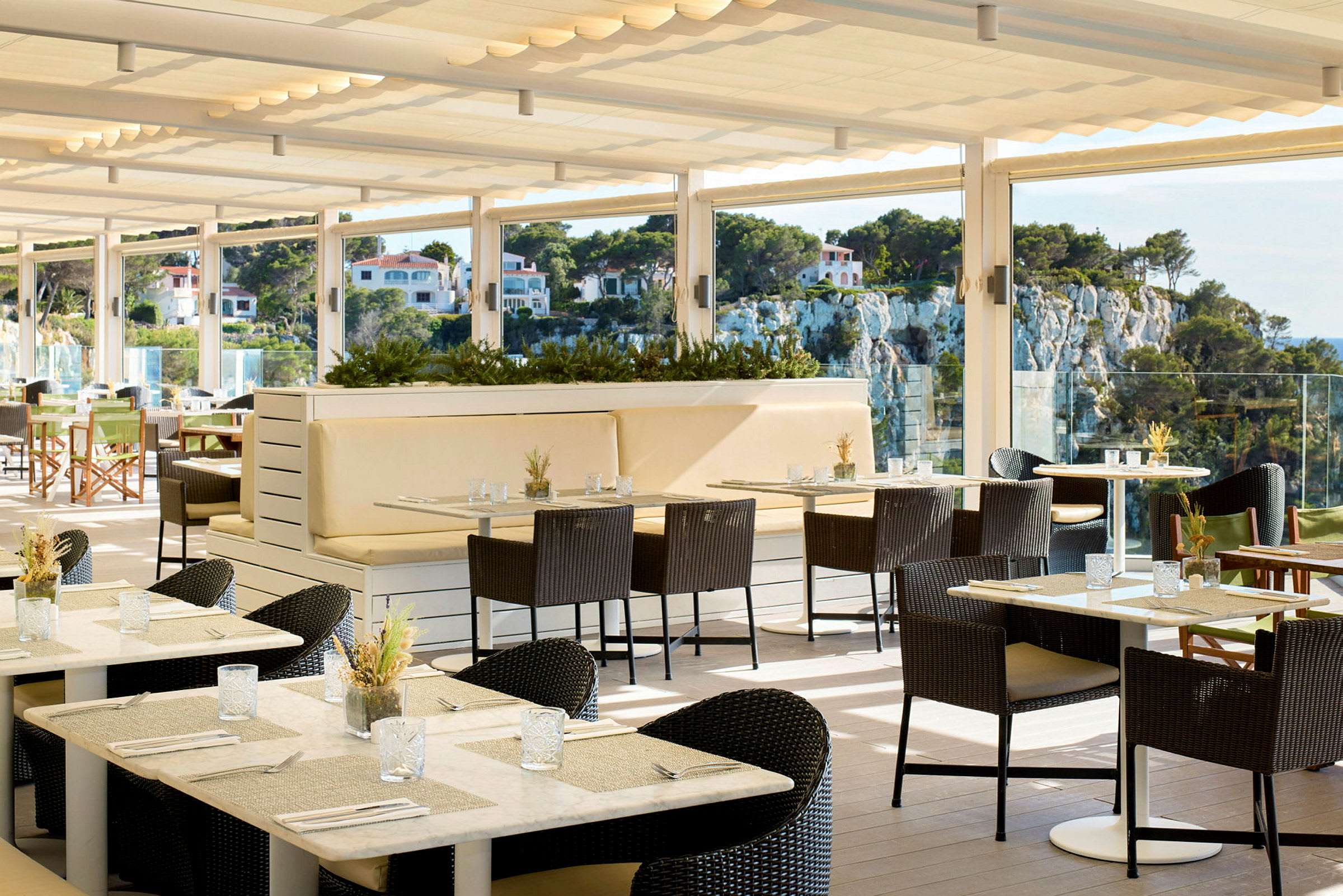 En solig restaurangterrass med bord och stolar samt utsikt över ett grönskande område med hus på en upphöjd klippa. (AI-genererad text)