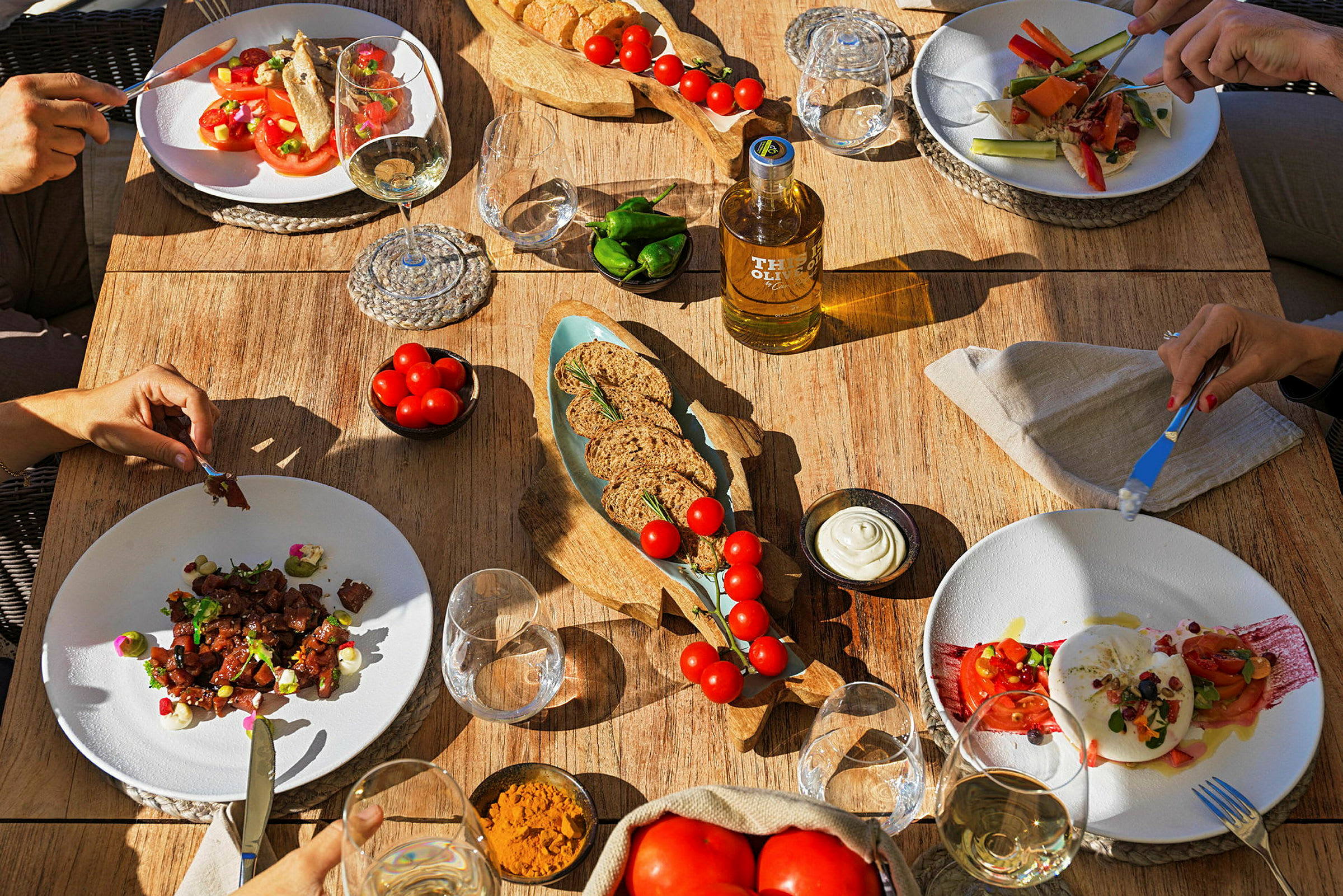 Ett matbord med tallrikar fyllda med sallad och andra rätter, omgivna av glas och skålar med olja, bröd och tillbehör. (AI-genererad text)