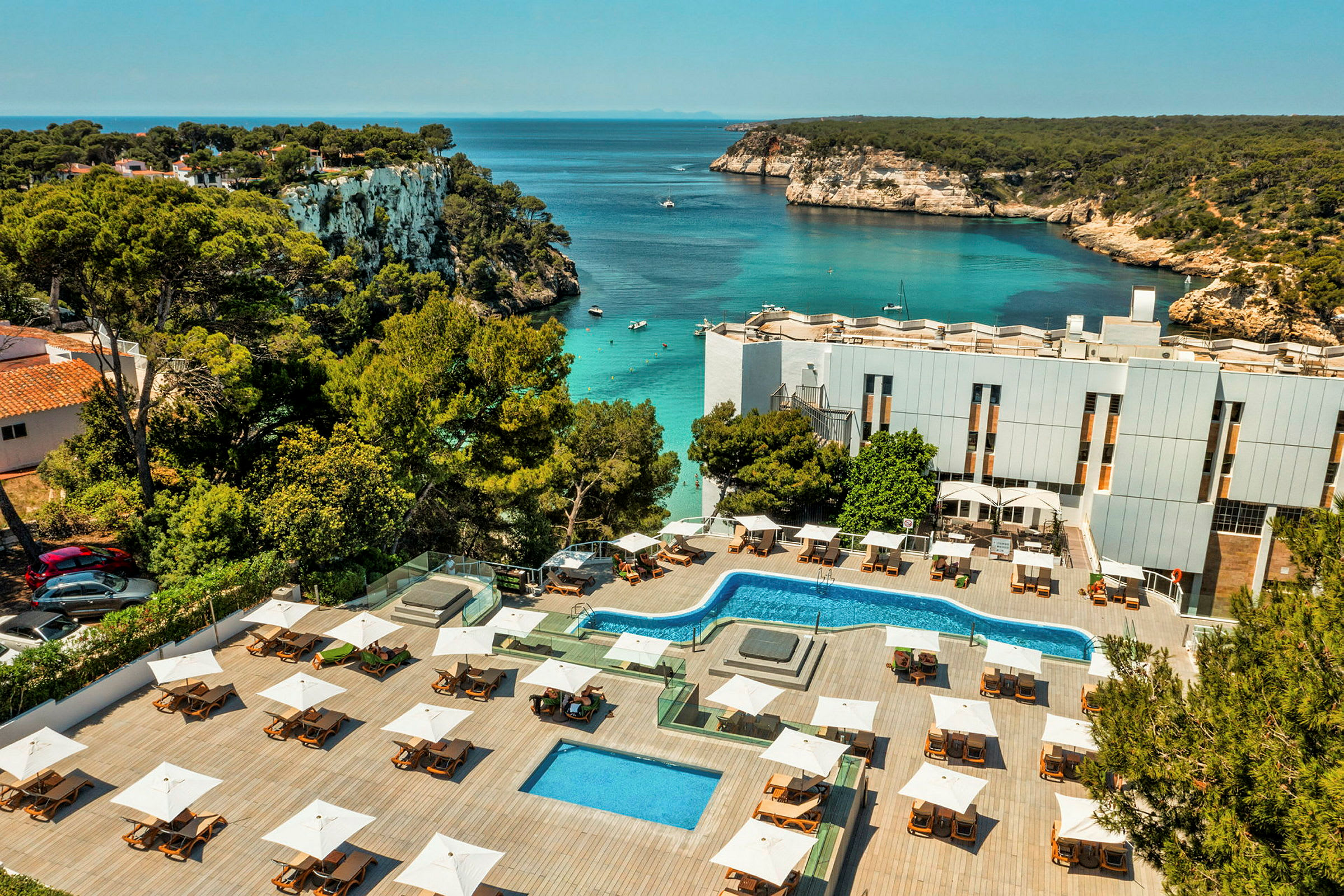 Ett hotellområde med pooler och solstolar ligger vid en vik med klart vatten och klippkust. (AI-genererad text)