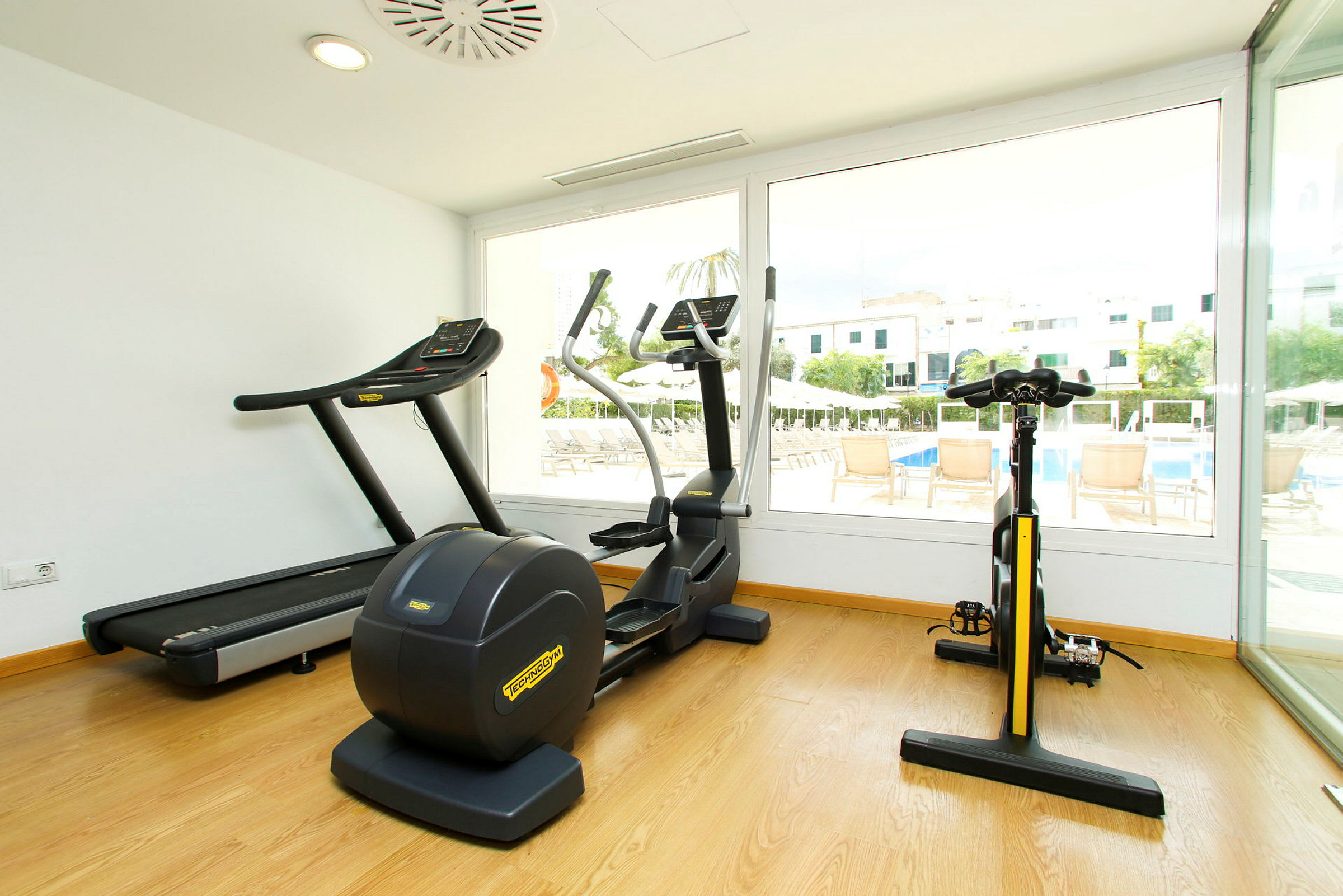 Ett gym med träningsmaskiner och stora fönster som vetter mot en pool och byggnader. (AI-genererad text)