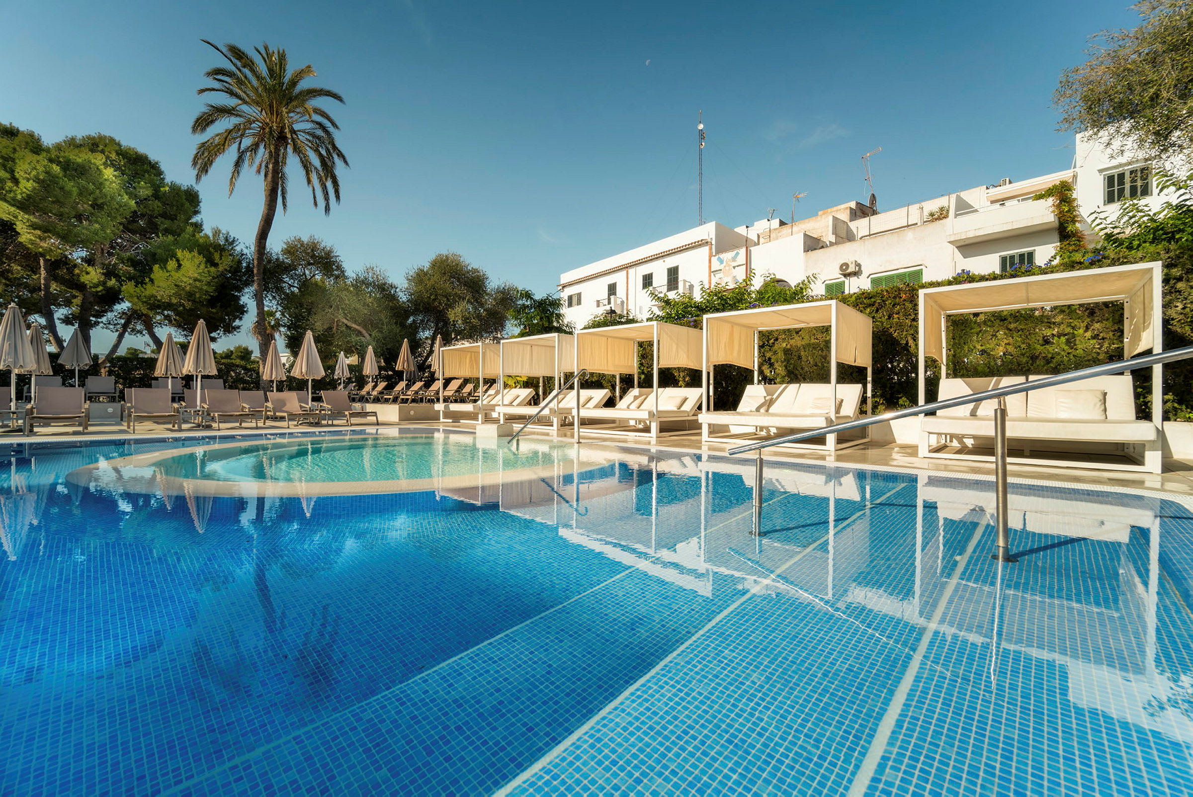 Bild av en pool med turkost vatten och vita solsängar under parasoll samt cabanas. Det är omgivande grönska och en skymt av byggnader i bakgrunden. (AI-genererad text)