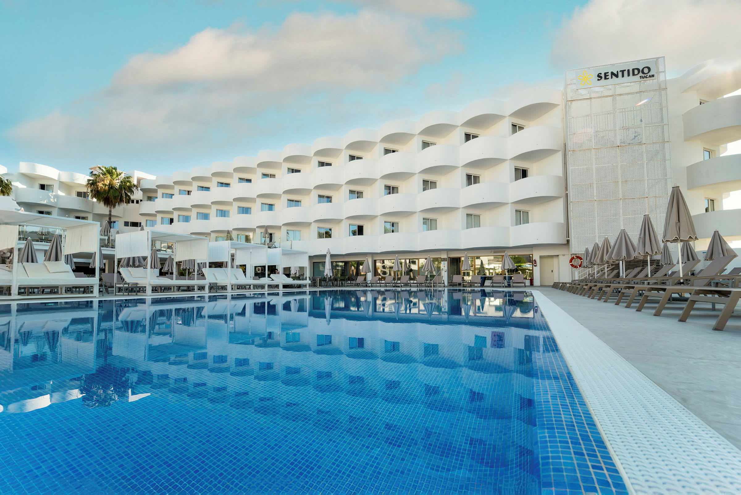 Bild av ett hotell med en stor pool framför. Byggnaden är vit med balkonger och solstolar vid poolområdet. (AI-genererad text)