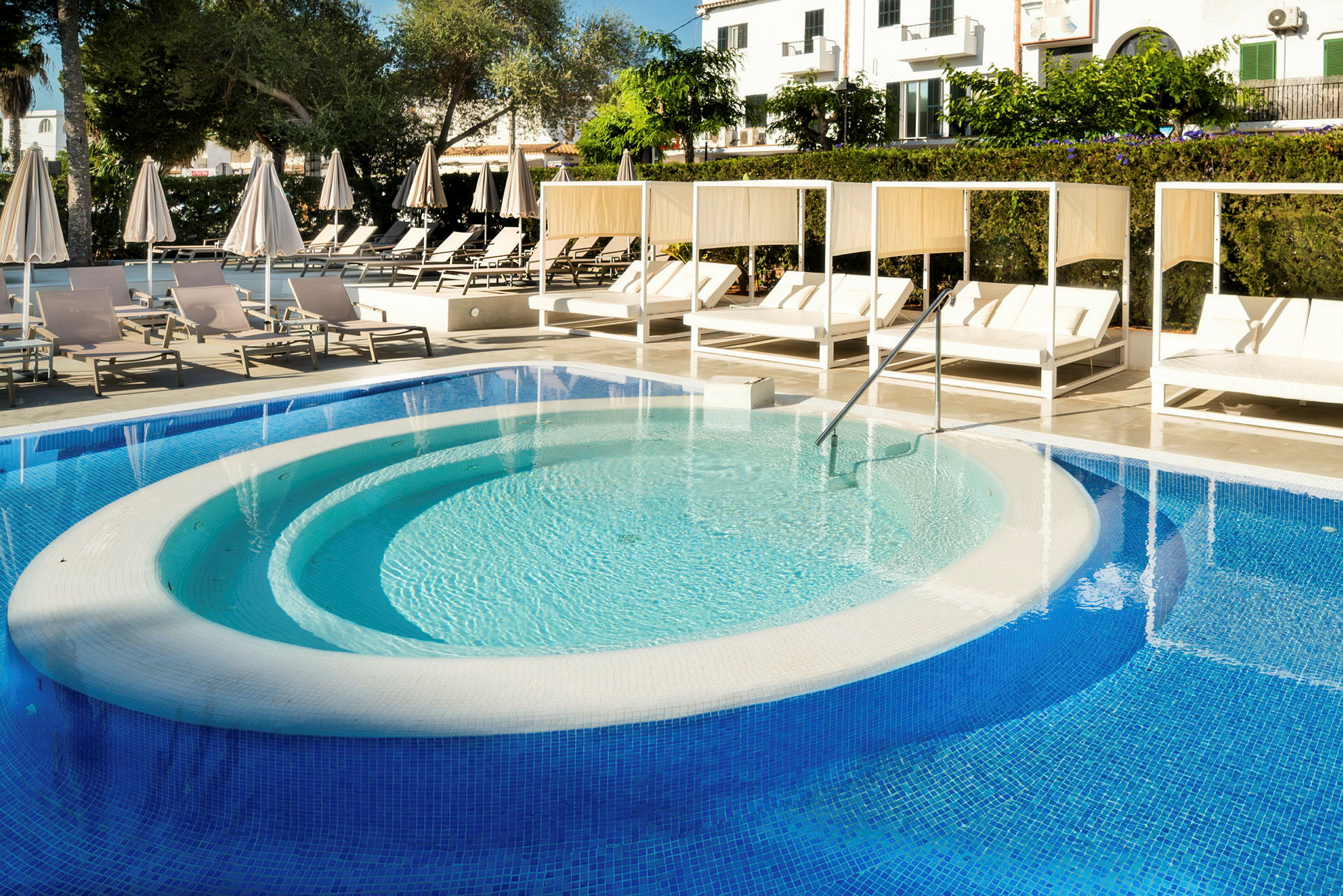 Bilden visar en swimmingpool med solstolar och parasoller runt omkring. Det finns även skuggade sittplatser vid kanten. (AI-genererad text)