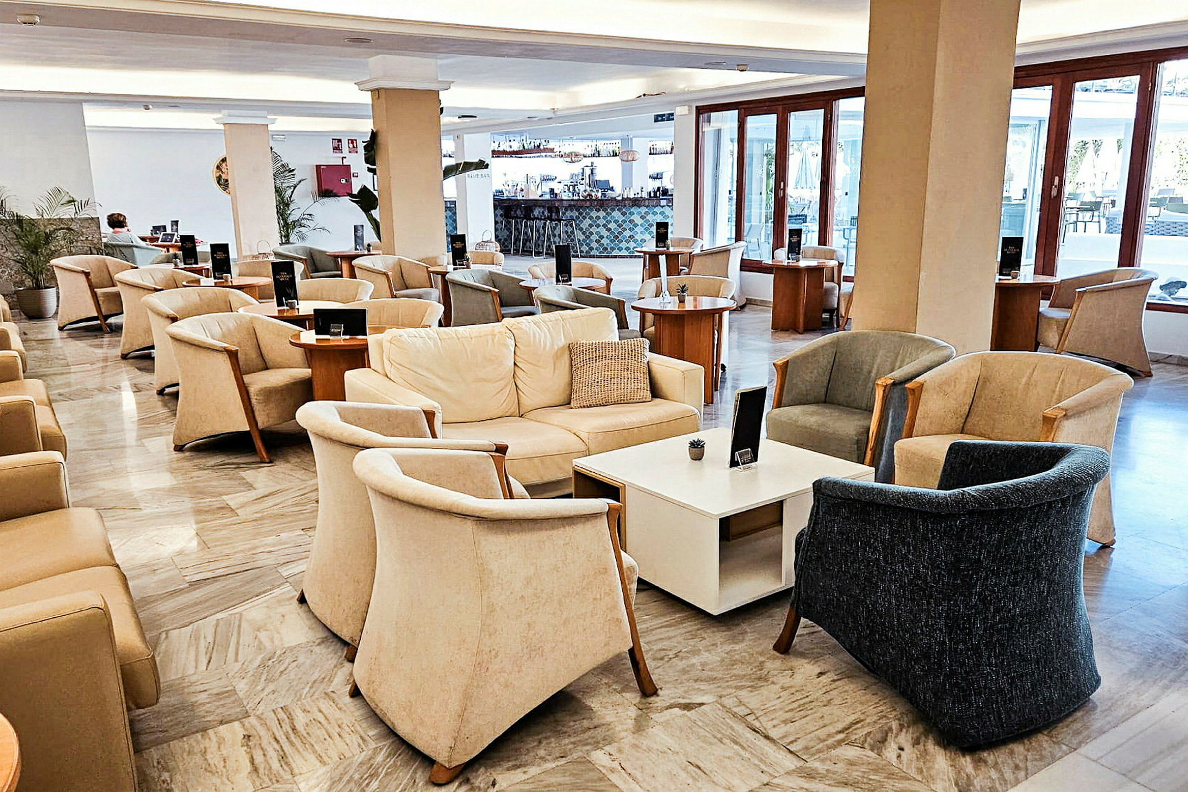 En lounge med bekväma fåtöljer, soffor och bord, med stora fönster i bakgrunden. (AI-genererad text)