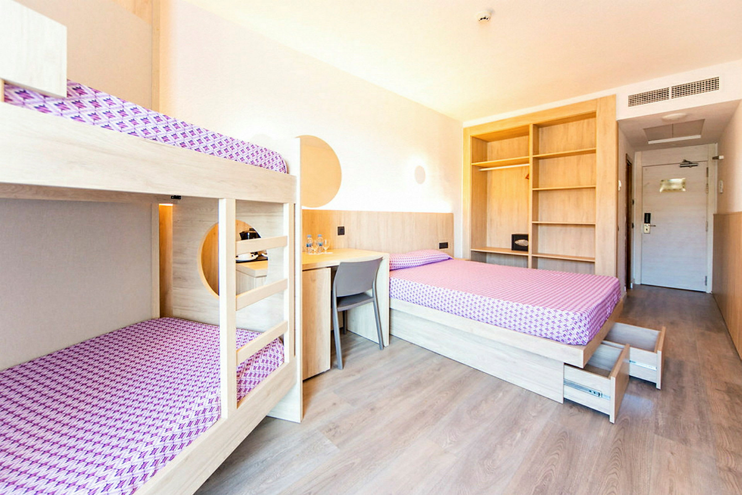 Ett hotellrum med våningssängar, en enkelsäng, skrivbord och öppna hyllor. Ett dörrområde kan ses i bakgrunden. (AI-genererad text)