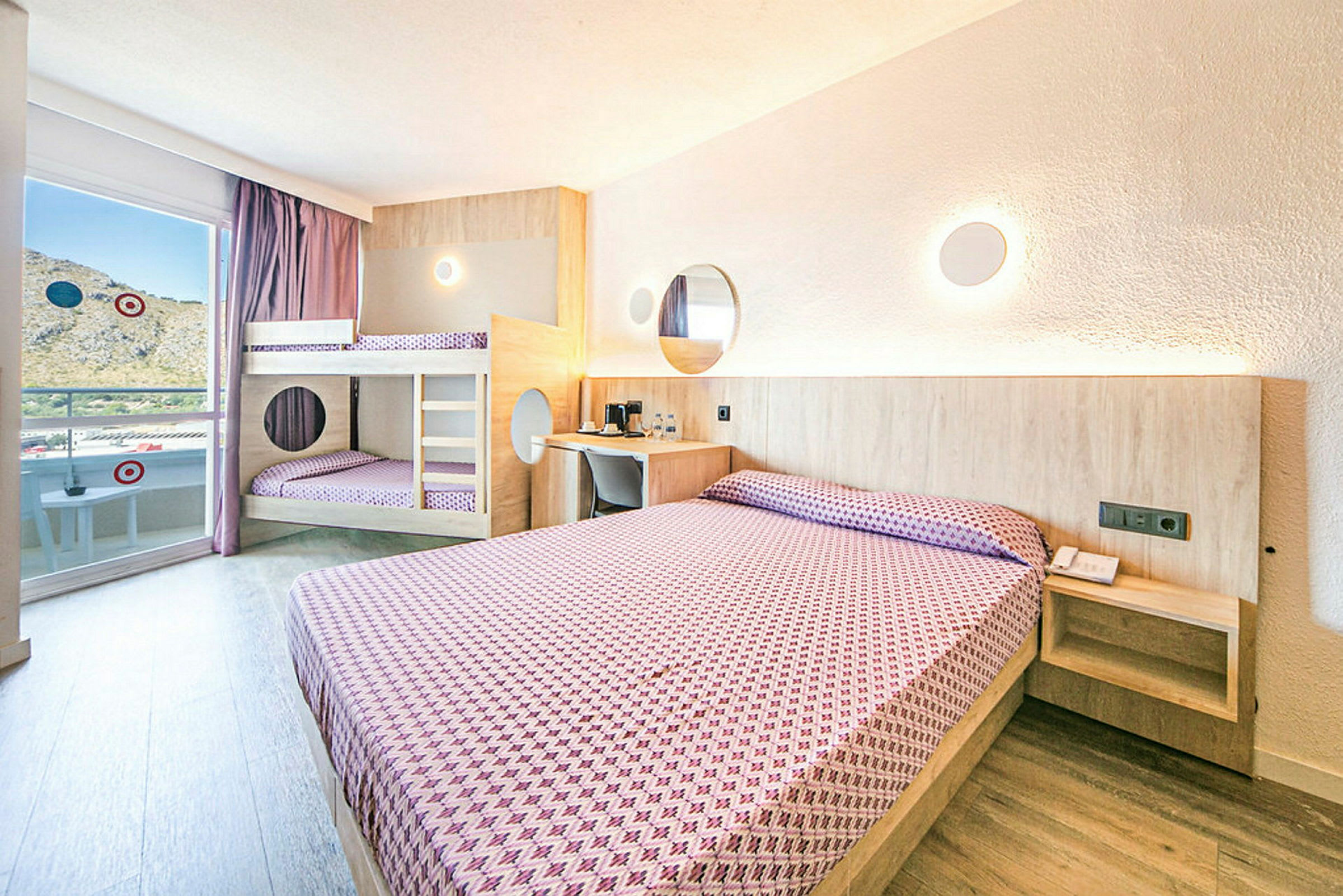 Ett hotellrum med en dubbelsäng och en våningssäng, båda bäddade med lila mönstrade sängkläder, ljusa väggar och trädetaljer. (AI-genererad text)