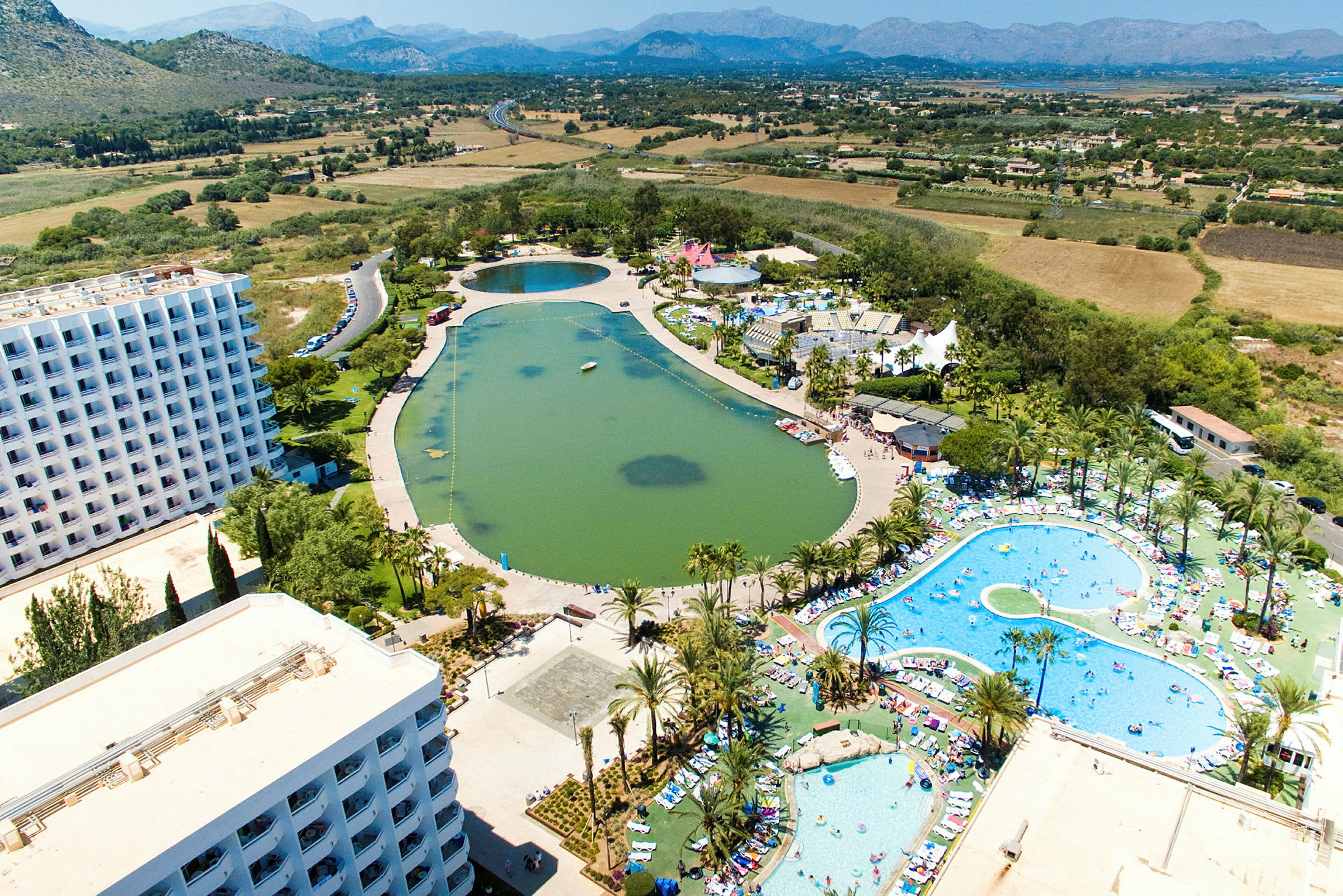 Flygbild av ett semesterkomplex med hotellbyggnader, pooler och en konstgjord sjö, omgivet av grönska och landskap. (AI-genererad text)