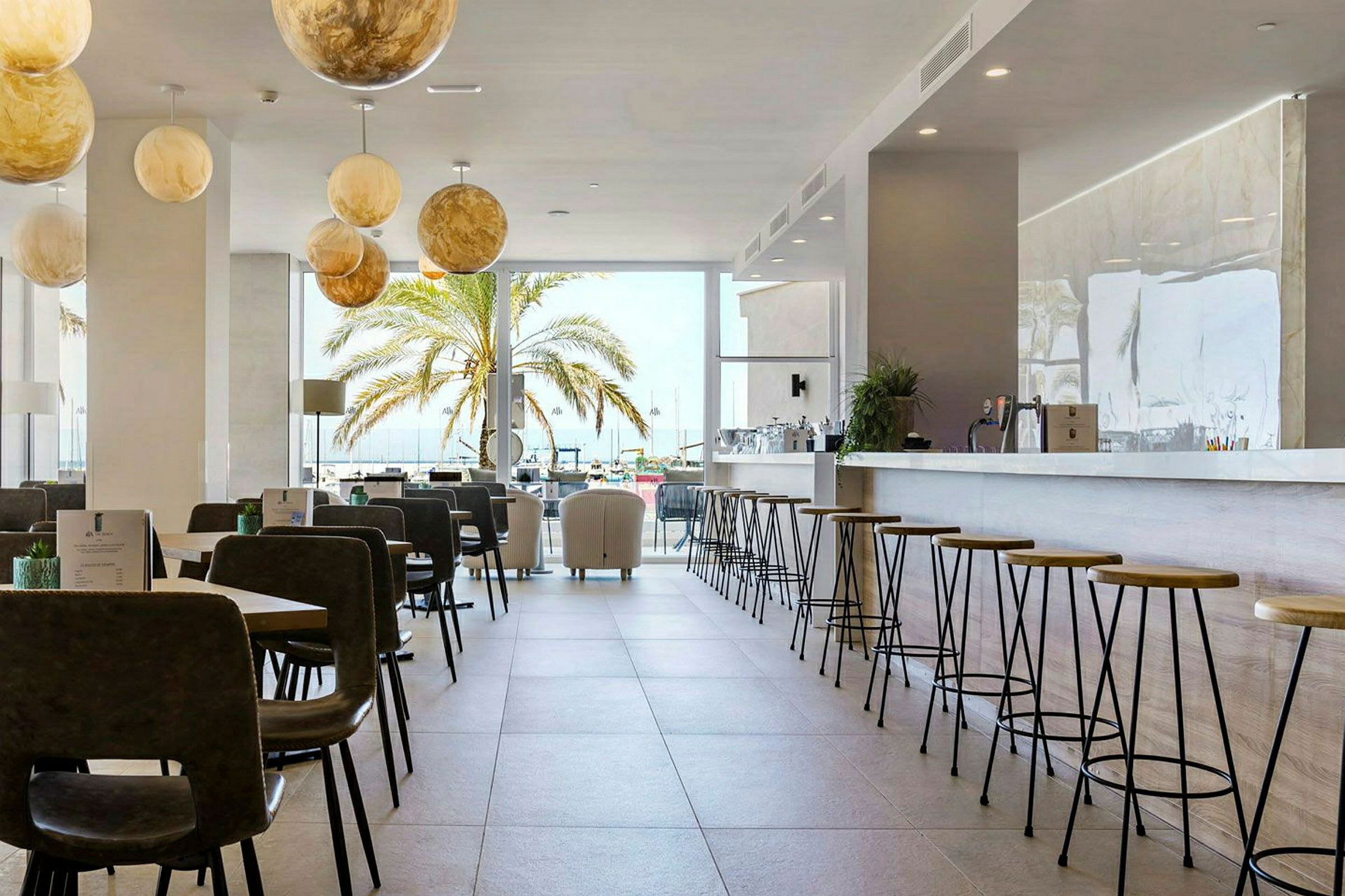 En ljus bar och restaurang med sittplatser, samt en palm utanför ett stort fönster. (AI-genererad text)