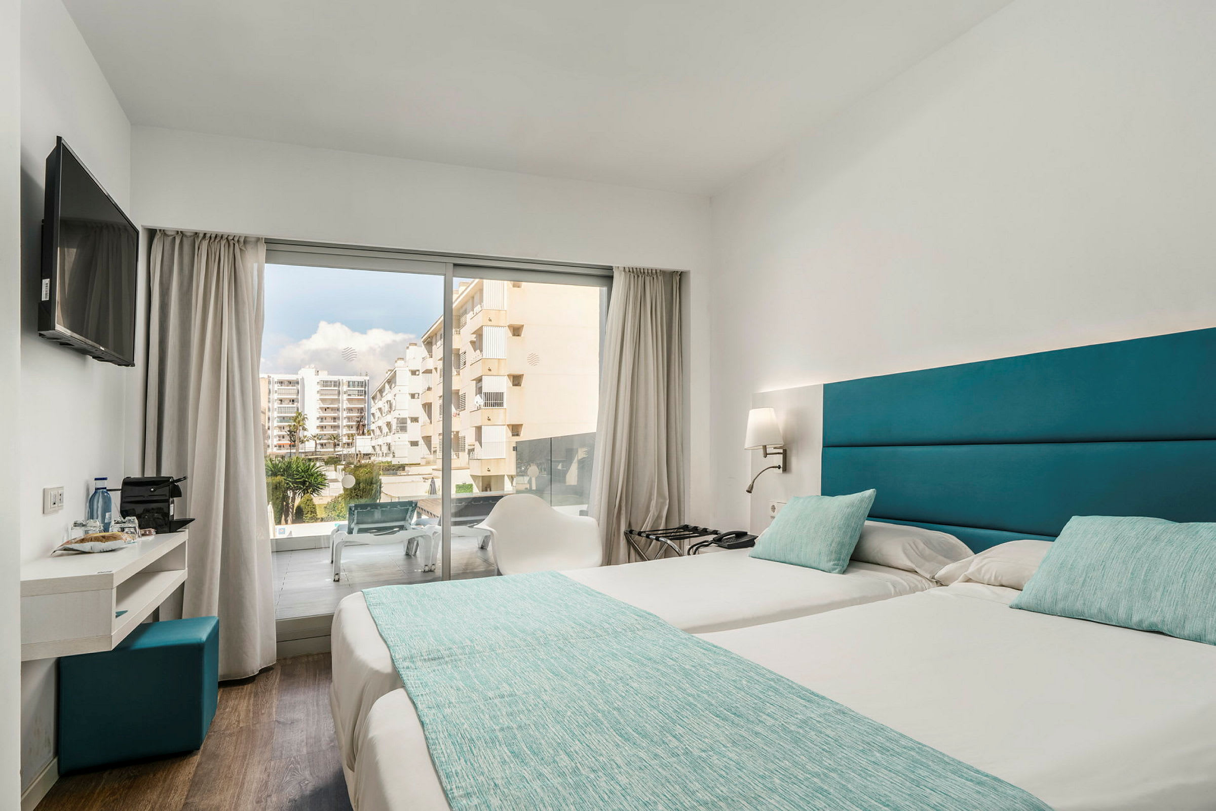 Bild av ett hotellrum med två enkelsängar bredvid varandra, turkosa sängkläder och kuddar samt en modern inredning. En glasdörr ger utsikt mot en balkong och ett område med byggnader. (AI-genererad text)