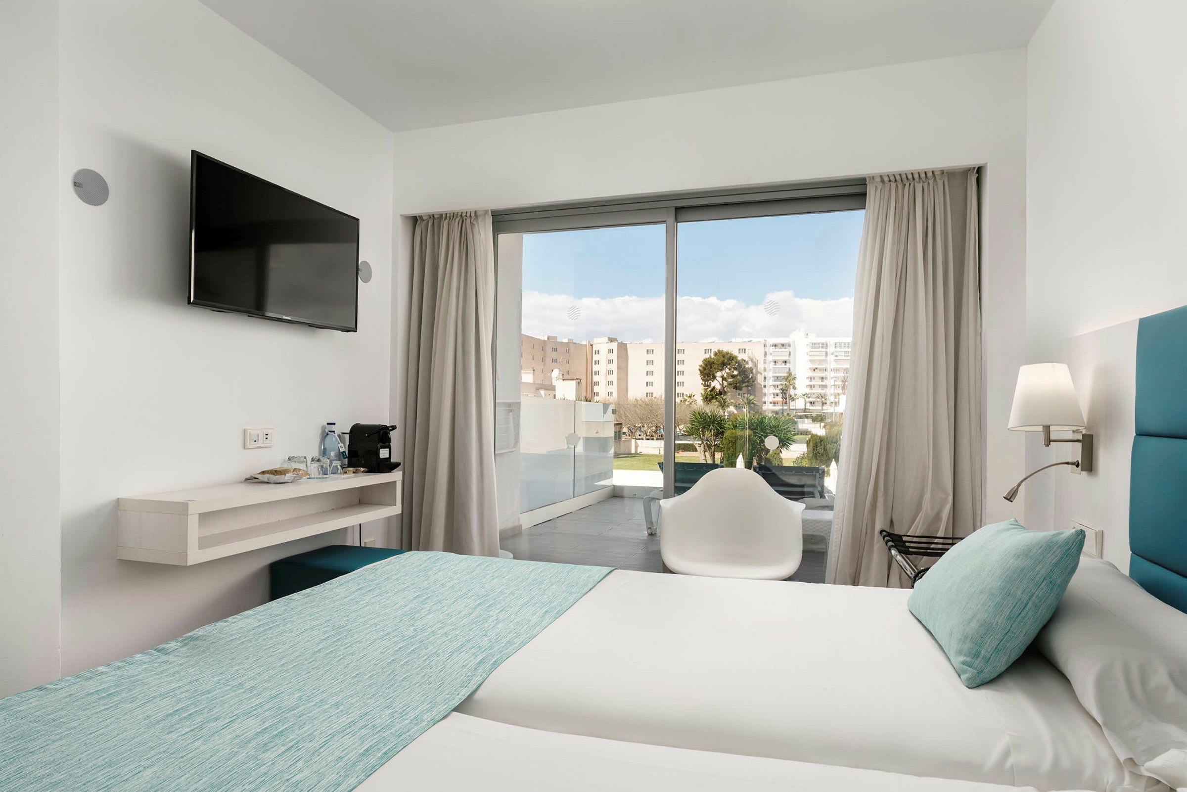 Ett hotellrum med två sängar bäddade med ljusa lakan och turkosa överkast. En stol står framför en glasdörr som leder ut till en balkong. En väggmonterad tv hänger bredvid en hylla med vattenflaskor och kaffemaskin. (AI-genererad text)