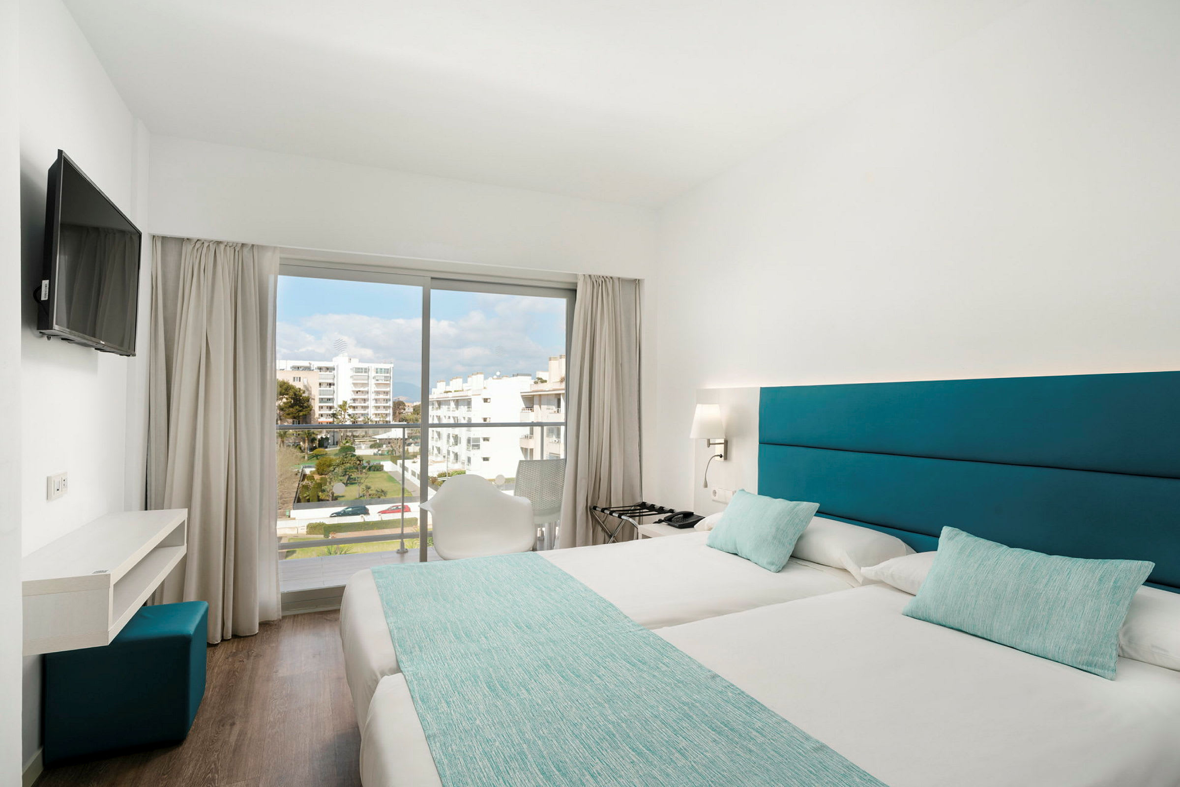 Ett hotellrum med två enkelsängar, en TV på väggen, ett skrivbord och en glasdörr som leder till en balkong med utsikt mot ett bostadsområde. (AI-genererad text)