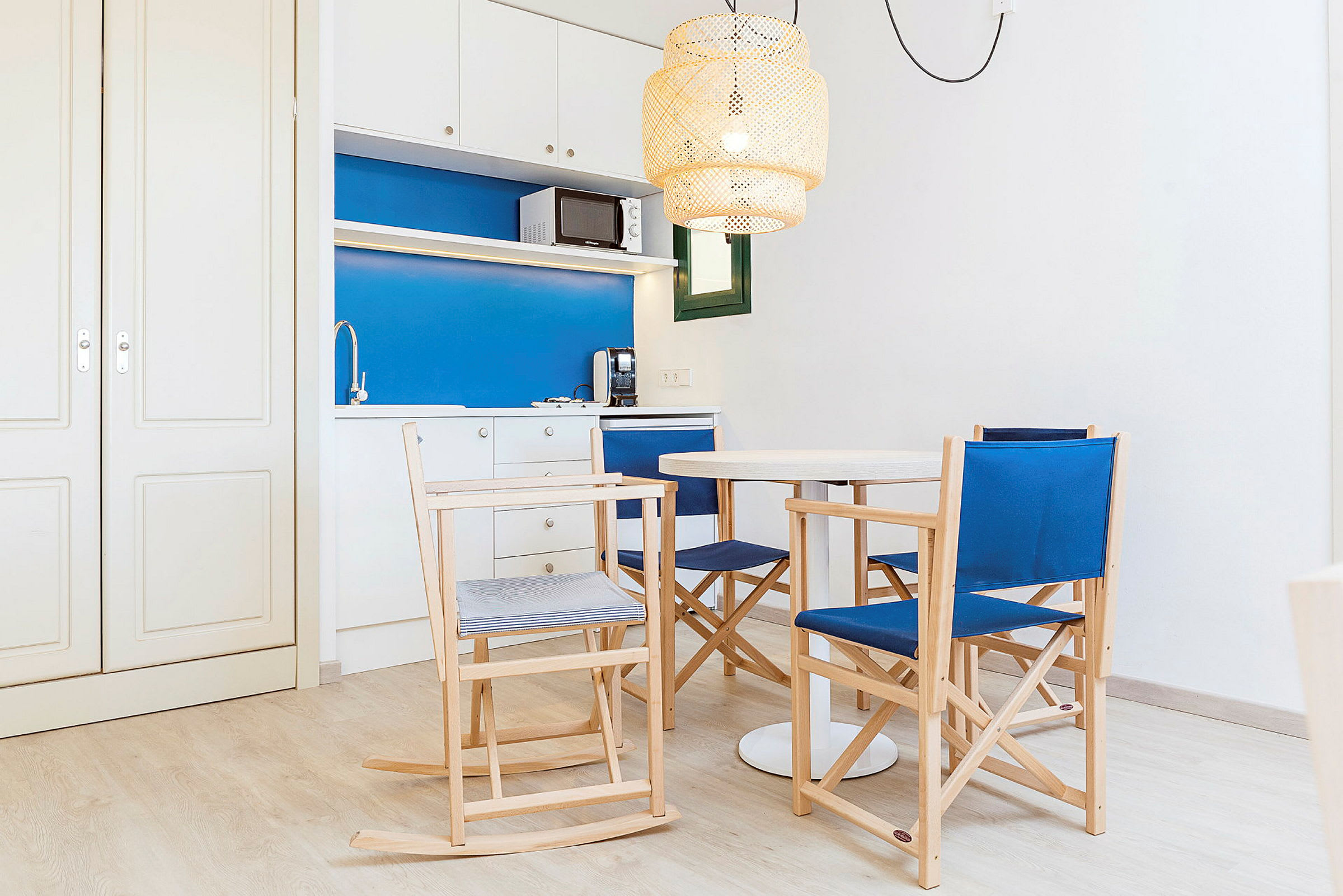 Ett litet kök med blå och vita ytor, en matgrupp med trästolar och en rund vit bord. En taklampa av flätad design hänger ovanför bordet. (AI-genererad text)