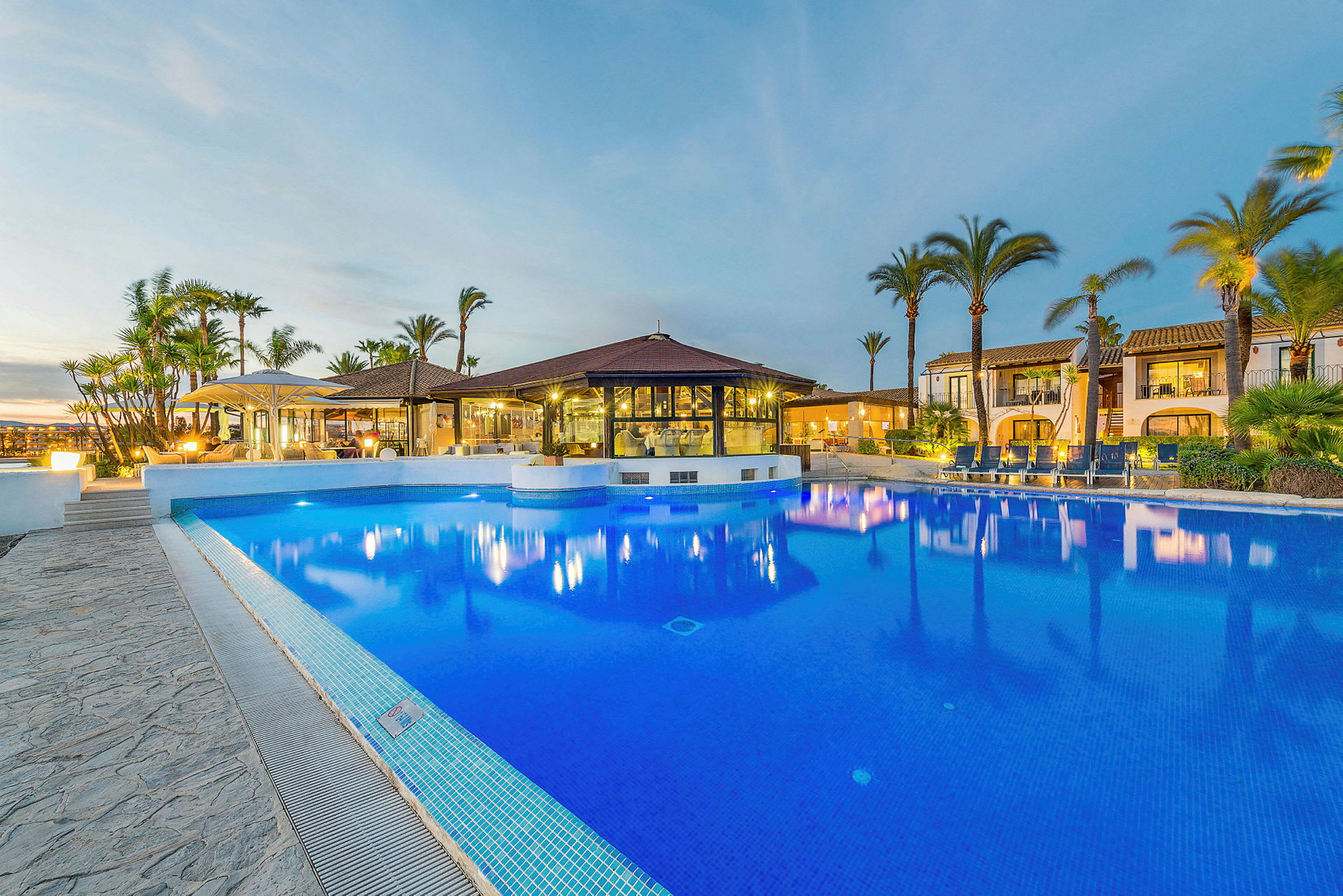 En pool vid ett hotell med palmer och byggnader i bakgrunden. (AI-genererad text)