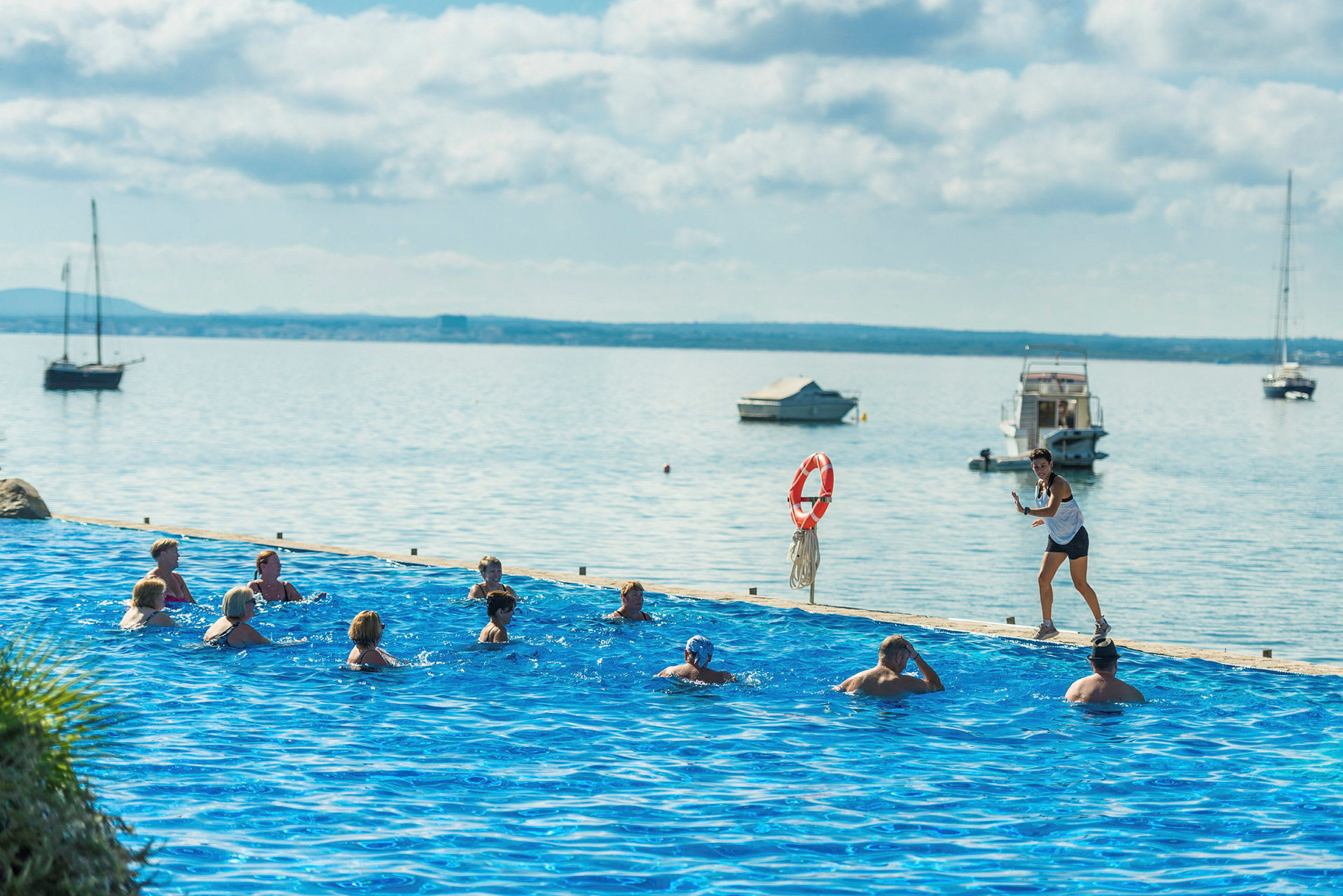 En grupp personer simmar i en pool nära vattnet med båtar i bakgrunden. (AI-genererad text)