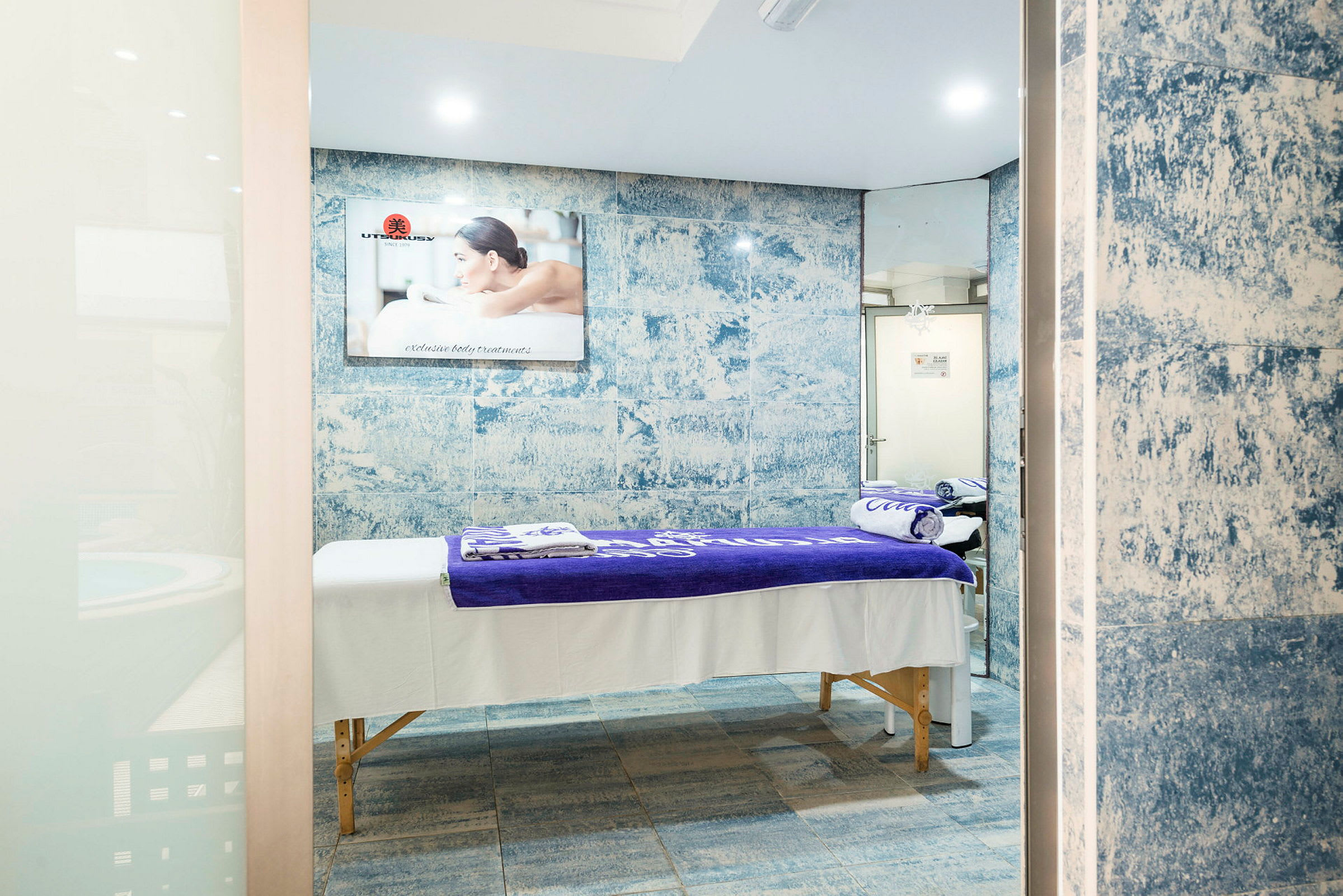 Ett spa-rum med massagebänk täckt av vita och lila handdukar, blå mosaik väggdesign och spegel. (AI-genererad text)