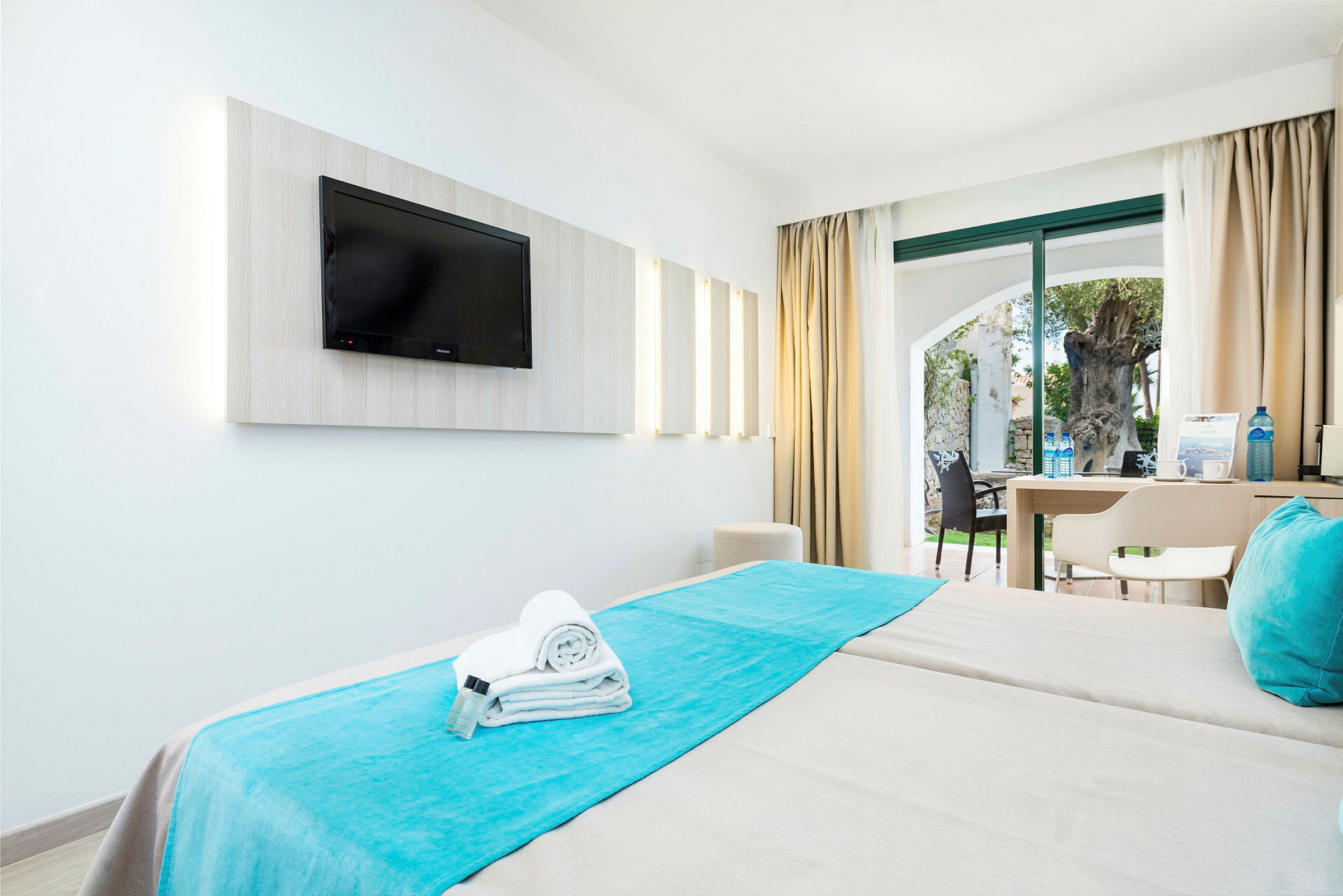Ett hotellrum med två sängar, en TV på väggen och en uteplats med utsikt över en trädgård genom stora fönster. (AI-genererad text)