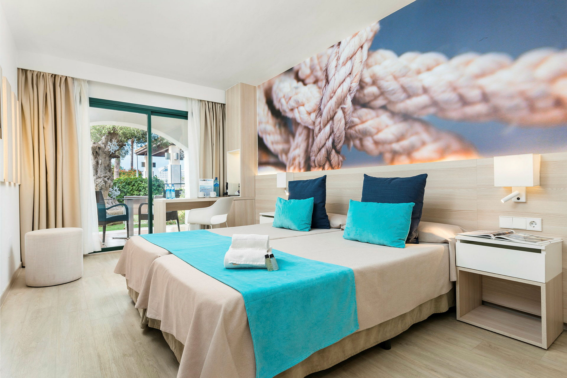 Modernt hotellrum med två sängar, blå detaljer och en bild av rep på väggen. (AI-genererad text)