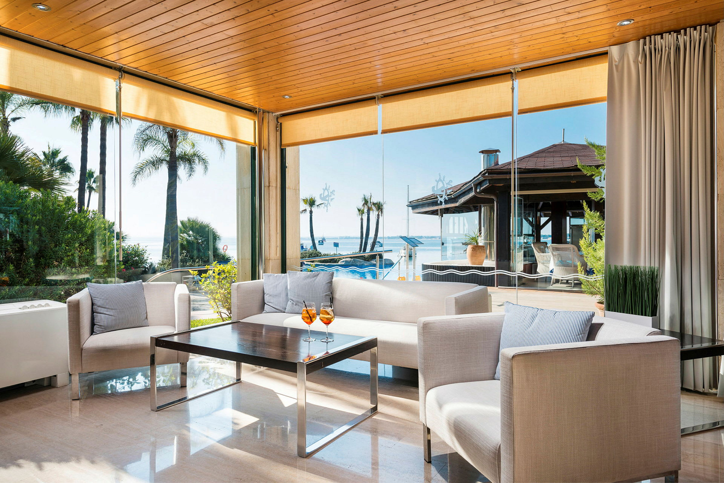 En ljus möblerad lounge med utsikt över havet genom stora glasfönster. (AI-genererad text)