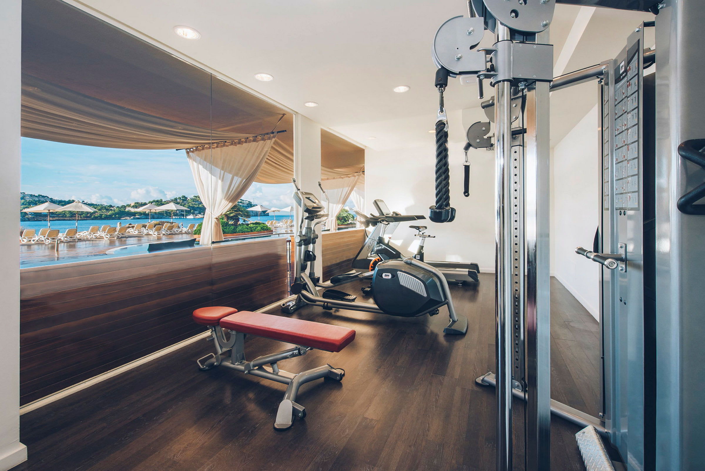 Ett gym med träningsmaskiner och en röd träningsbänk, med utsikt mot en pool och solstolar genom stora glasfönster. (AI-genererad text)