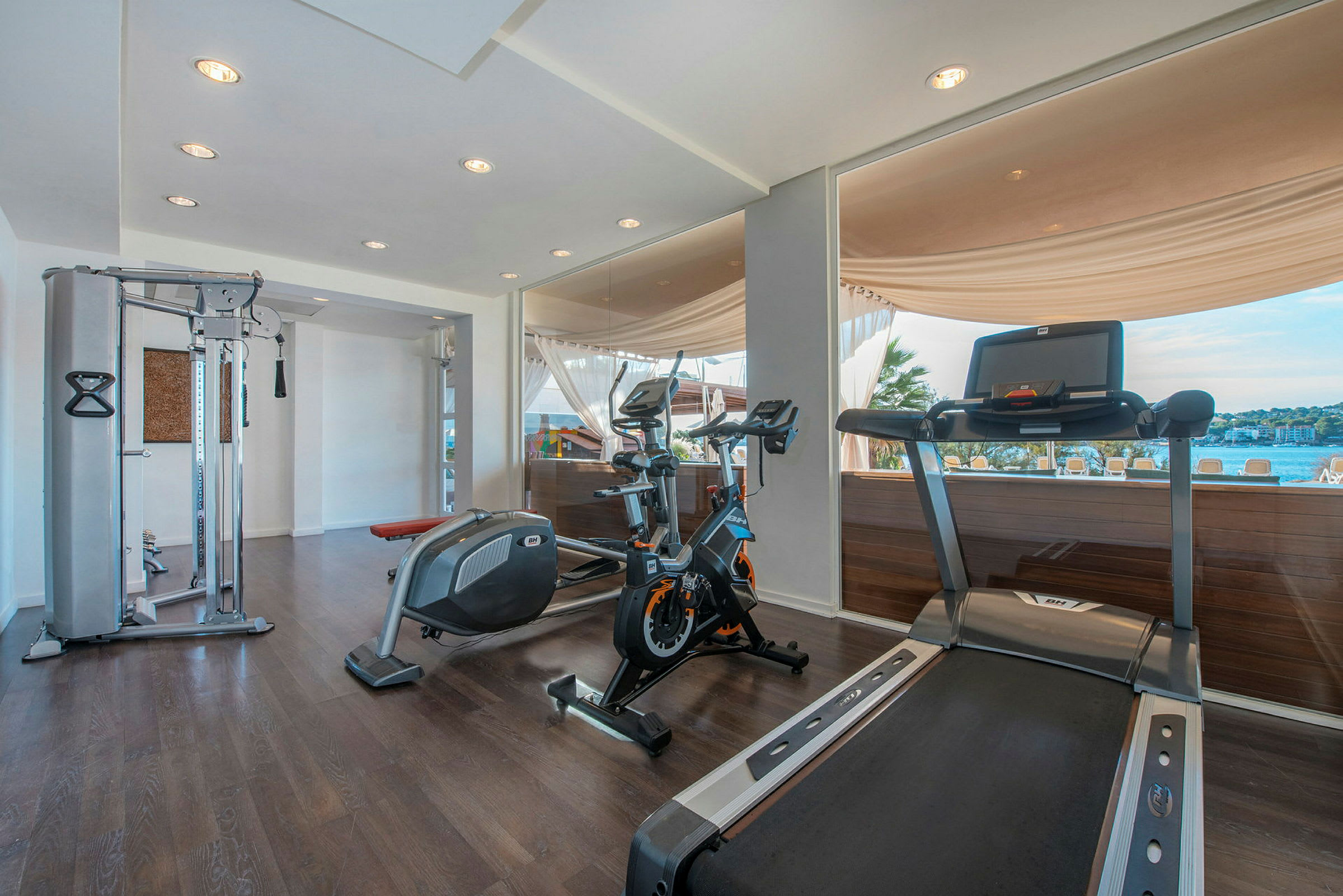 Ett gym med träningsutrustning som träningscykel, löpband och multifunktionsmaskin, med stora fönster som erbjuder utsikt över en pool och landskap utanför. (AI-genererad text)