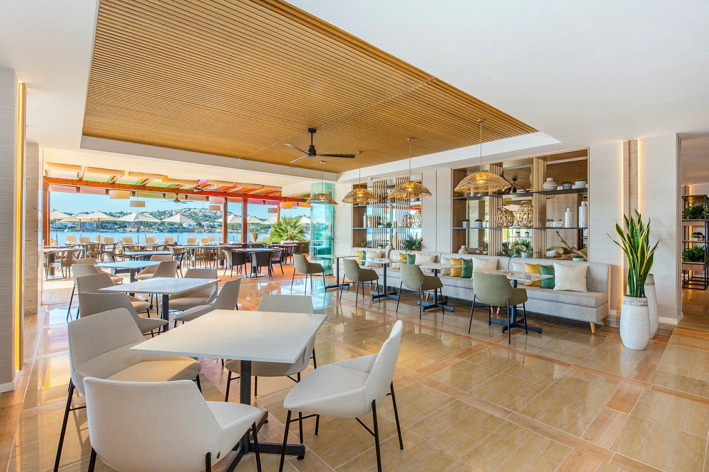 Ett modernt och öppet restaurangområde med bord och stolar, inrett med ljusa färger och stora fönster som vetter ut mot en utomhusterrass och utsikt över havet. (AI-genererad text)