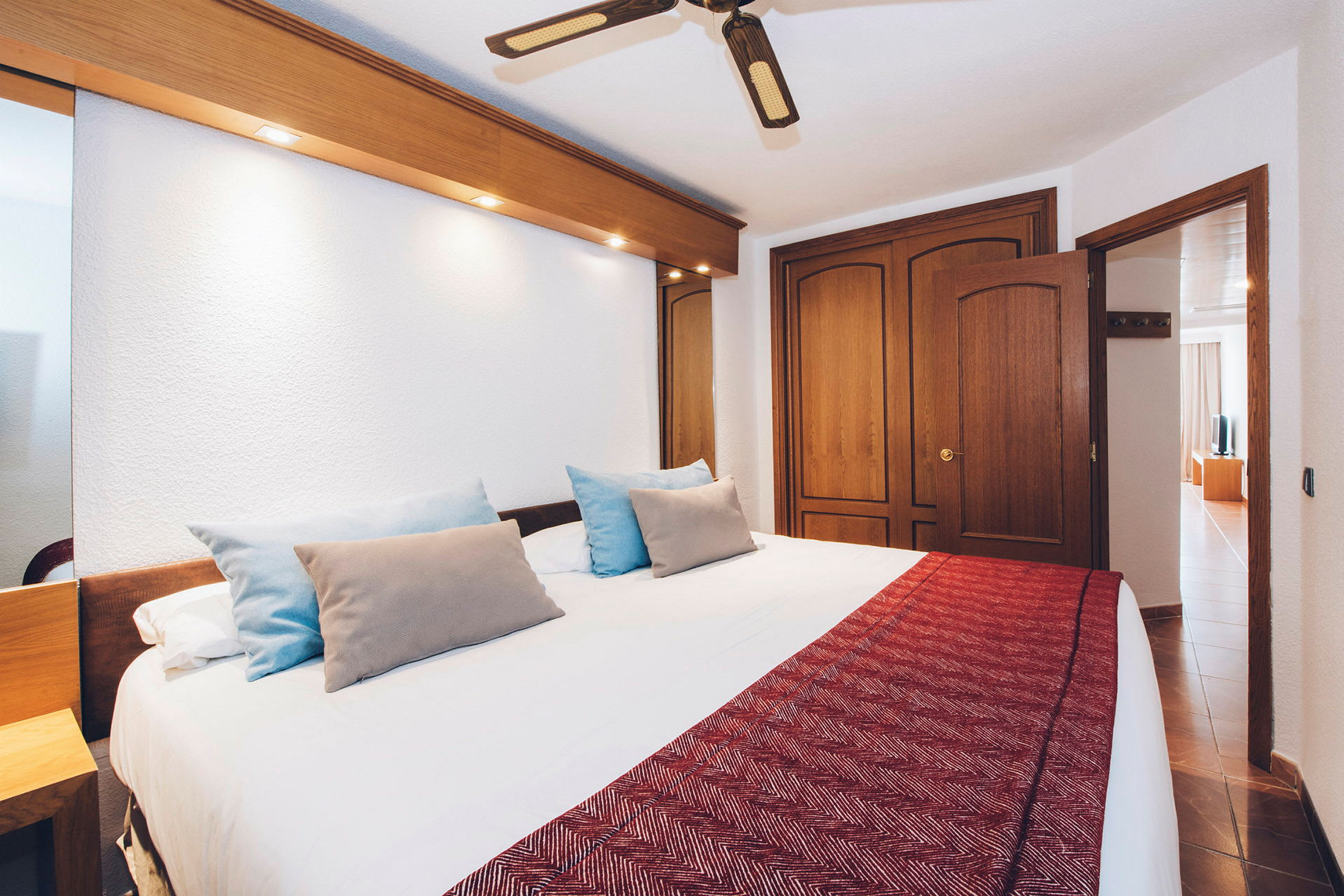 Ett hotellrum med en stor säng med kuddar och en röd sängfilt. Rummet har en takfläkt och en garderob med trädörrar. (AI-genererad text)