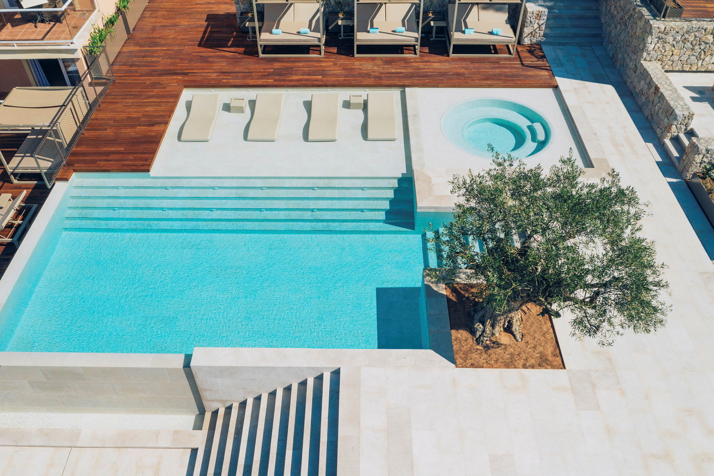 En modern pool med närliggande jacuzzi och träaltan. På altanen står några solsängar och det finns ett stort olivträd planterat vid poolkanten. (AI-genererad text)