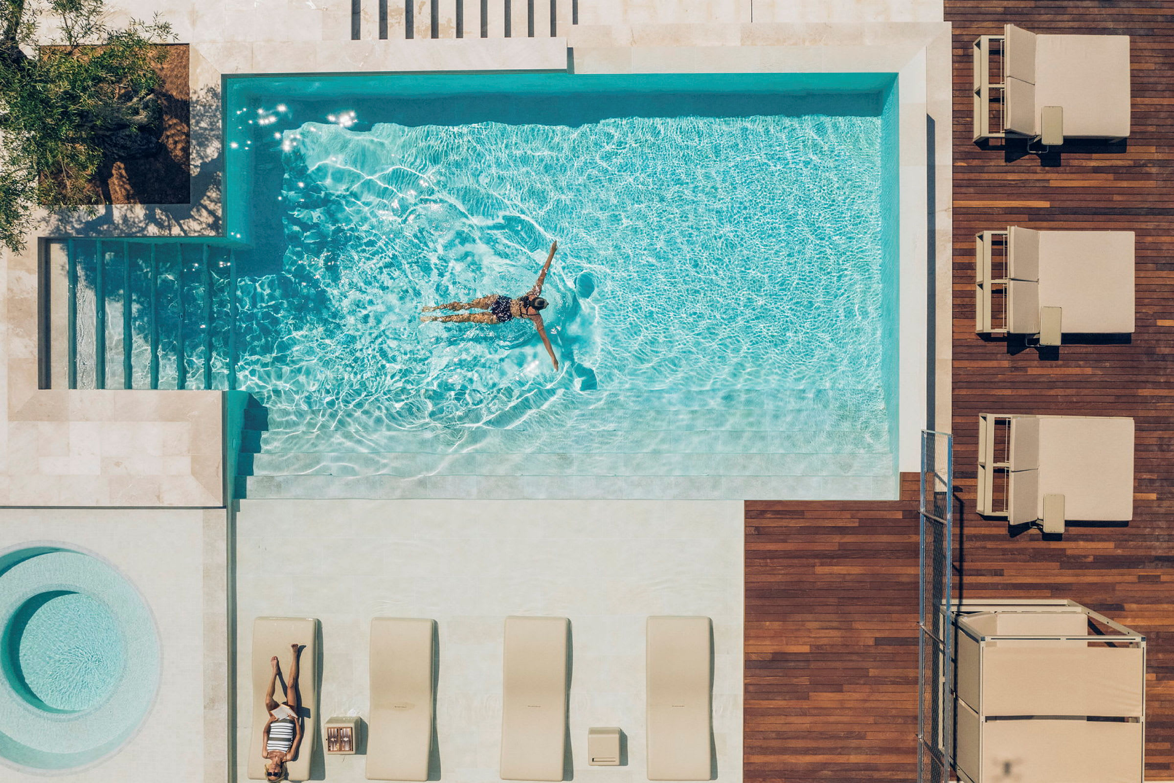 Bilden visar en översikt över en uteplats med en pool. I poolen simmar en person medan andra solstolar är placerade bredvid på trädäcket. (AI-genererad text)